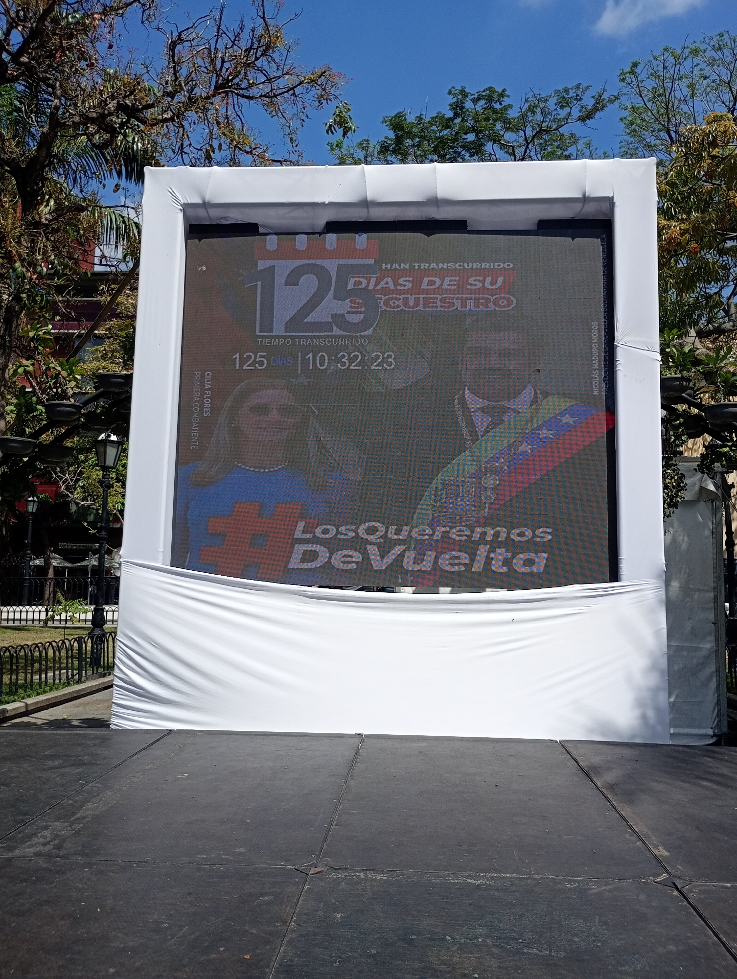 The image shows a large digital billboard on a stage in an outdoor area, likely a public space. The billboard displays a message related to a kidnapping, showing pictures of Cilia Flores (identified as "Primera Combatiente") and Nicolas Maduro (identified as "Presidente de la Republica Bolivariana de Venezuela"). Text on the screen says "125 Dias de Su Cuestro" (125 Days of Their Kidnapping), with a time elapsed of 125 days and 10 hours, 32 minutes, and 23 seconds. The phrase "Los Queremos Devuelta" (We Want Them Back) is prominently displayed. The billboard is surrounded by a white frame and draped with white fabric at the bottom. In the background are trees and a black wrought-iron fence, suggesting a park or plaza. The photo was likely taken during the day, given the bright sunlight. The location is suggested to be in Caracas, Venezuela.