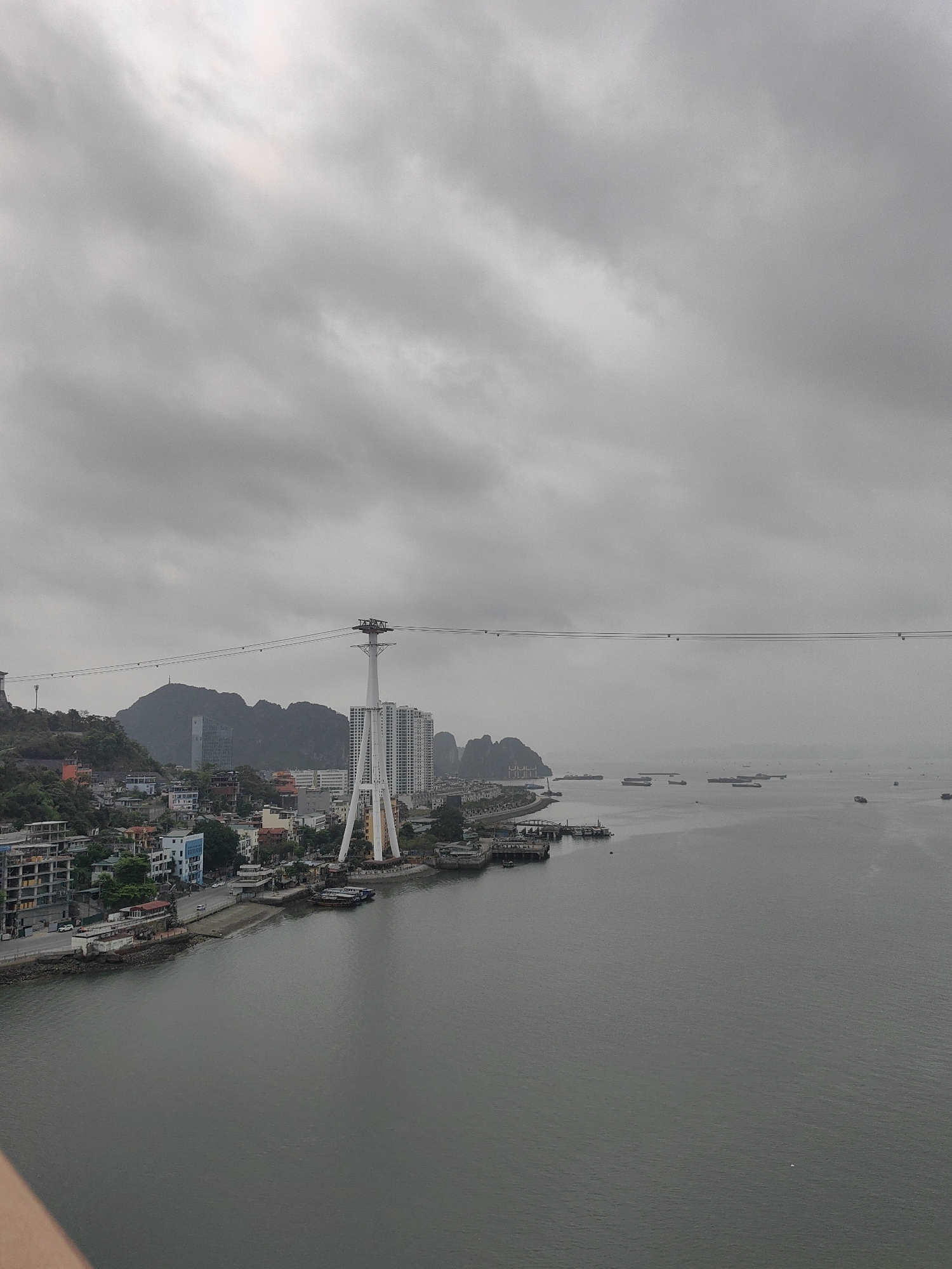 This image depicts a section of Hong Gai Ward, Vietnam, under a cloudy sky. The view encompasses a waterfront area with a mix of residential and commercial buildings, situated along a bay or inlet. A large, white cable car tower stands prominently, with cables stretching across the frame towards the horizon. The water contains several boats and floating structures. Mountains are visible in the background. The overall color palette is muted, dominated by gray tones due to the overcast weather.