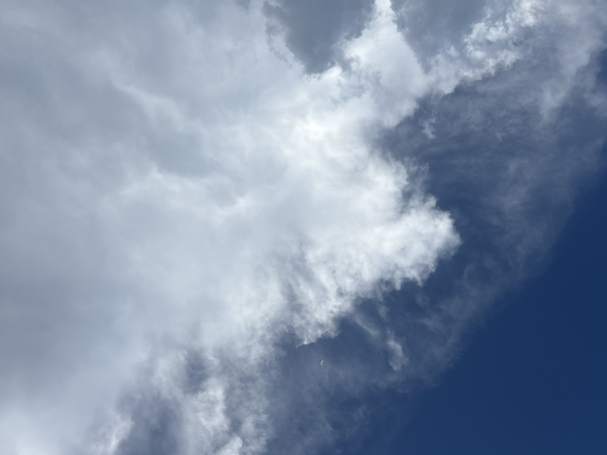 The image shows a daytime sky with clouds. The left side of the image is filled with thick, white clouds. The right side of the image is a clear, deep blue sky. There is a crescent moon visible near the lower center of the image, embedded within a cluster of thin clouds. The location is unknown and there is nothing to indicate that it is the city of Bukuru, Nigeria. There are no people, subjects, objects, actions, or interactions present. The environment consists of the sky and clouds.