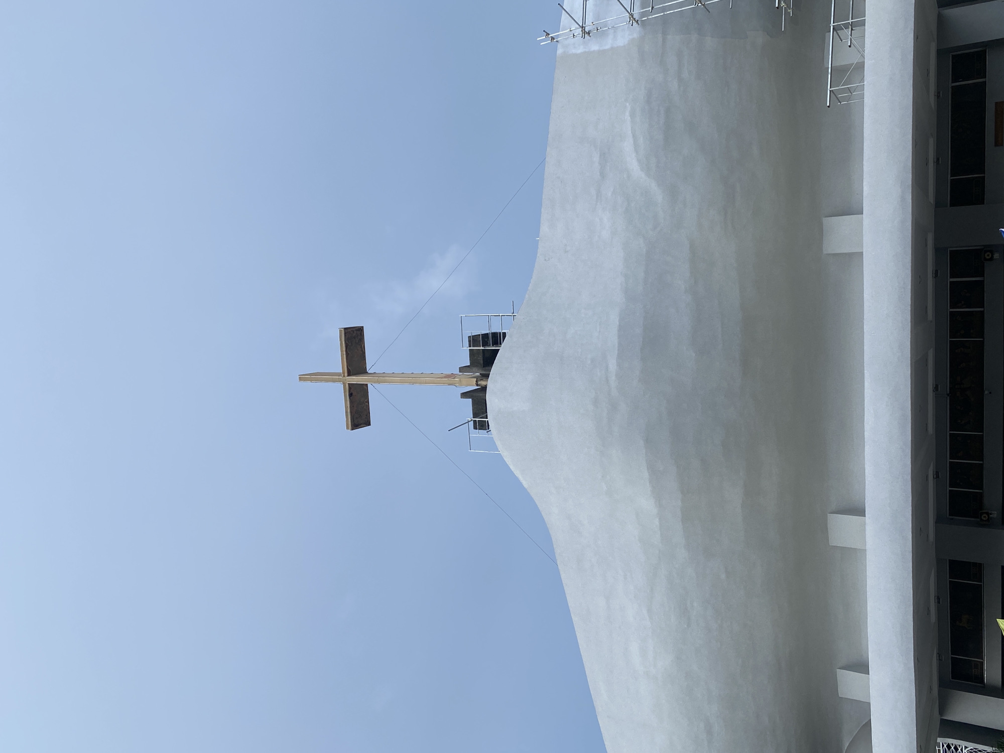 This image shows the exterior of a building with a large, light gray, undulating roof. At the apex of the roof, a wooden cross is mounted. The cross is simple in design, with a horizontal beam atop a vertical beam. The building appears to be under construction or renovation, as indicated by scaffolding on the right side of the structure and some exposed construction elements around the base of the cross. The sky above is overcast with a pale blue hue. Below the undulating roof, there is a section with a flat overhang supported by square columns, beneath which windows or display areas are visible. This building is the Parish of the Risen Lord, located along Mac Arthur Highway, Barangay Malagasang 1-D, Imus City, Cavite. However, the provided description of the location is not accurate as this building is not in Davao City, Philippines.