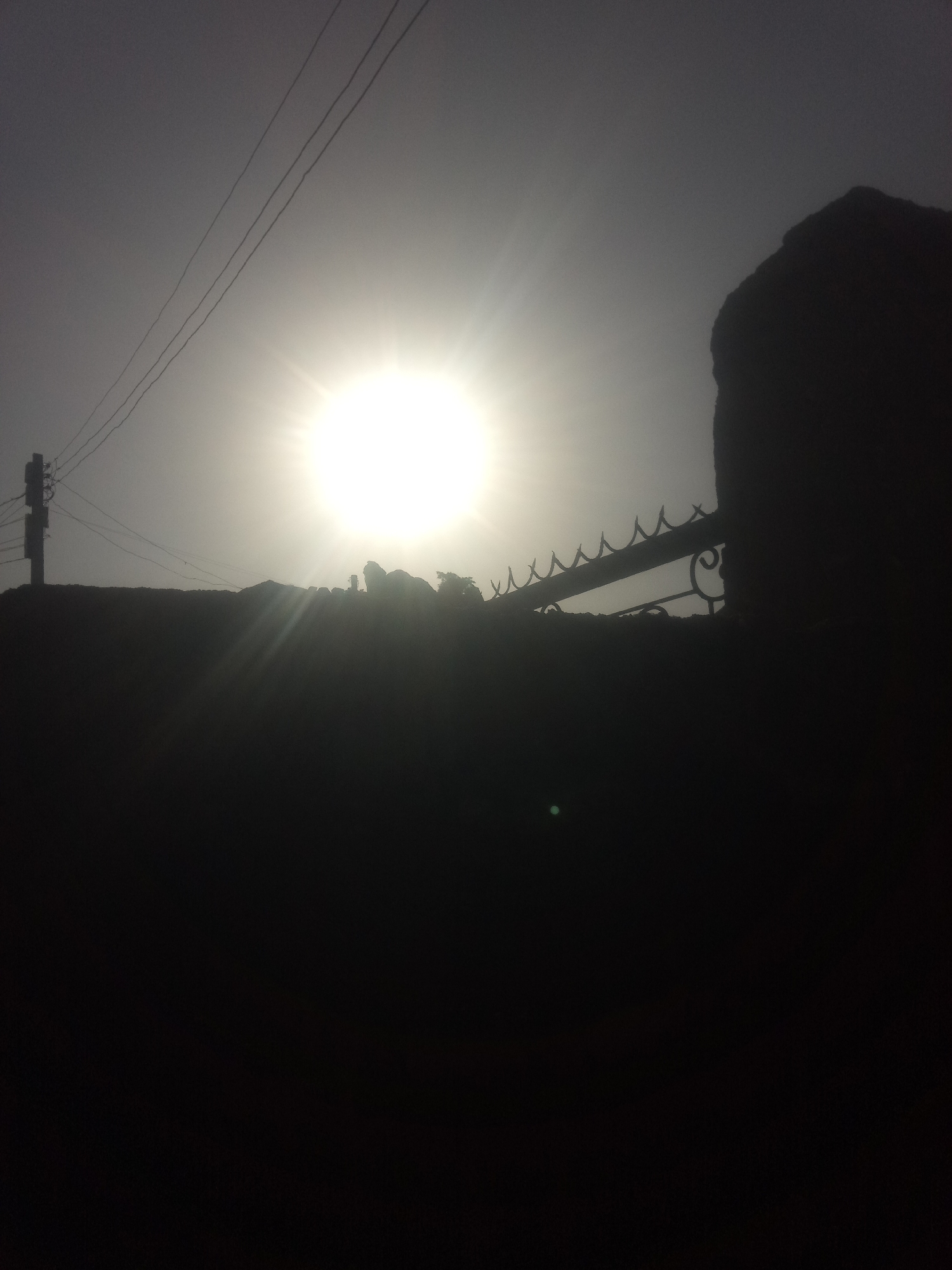 A silhouette of a fence and a wall against a bright sun is shown in the image. The sun is in the upper center of the image, with its rays emanating outwards. The fence appears to be made of metal with decorative elements. A portion of a dark, solid wall is on the right side of the image, partially obscuring the sun. The foreground is also in silhouette, showing what appears to be a raised surface or ground level. The sky is hazy or overcast, which diffuses the sunlight. A utility pole with wires is visible on the left side of the image, also in silhouette. The overall impression is of a bright, possibly dusty or hazy day, with strong backlighting from the sun.