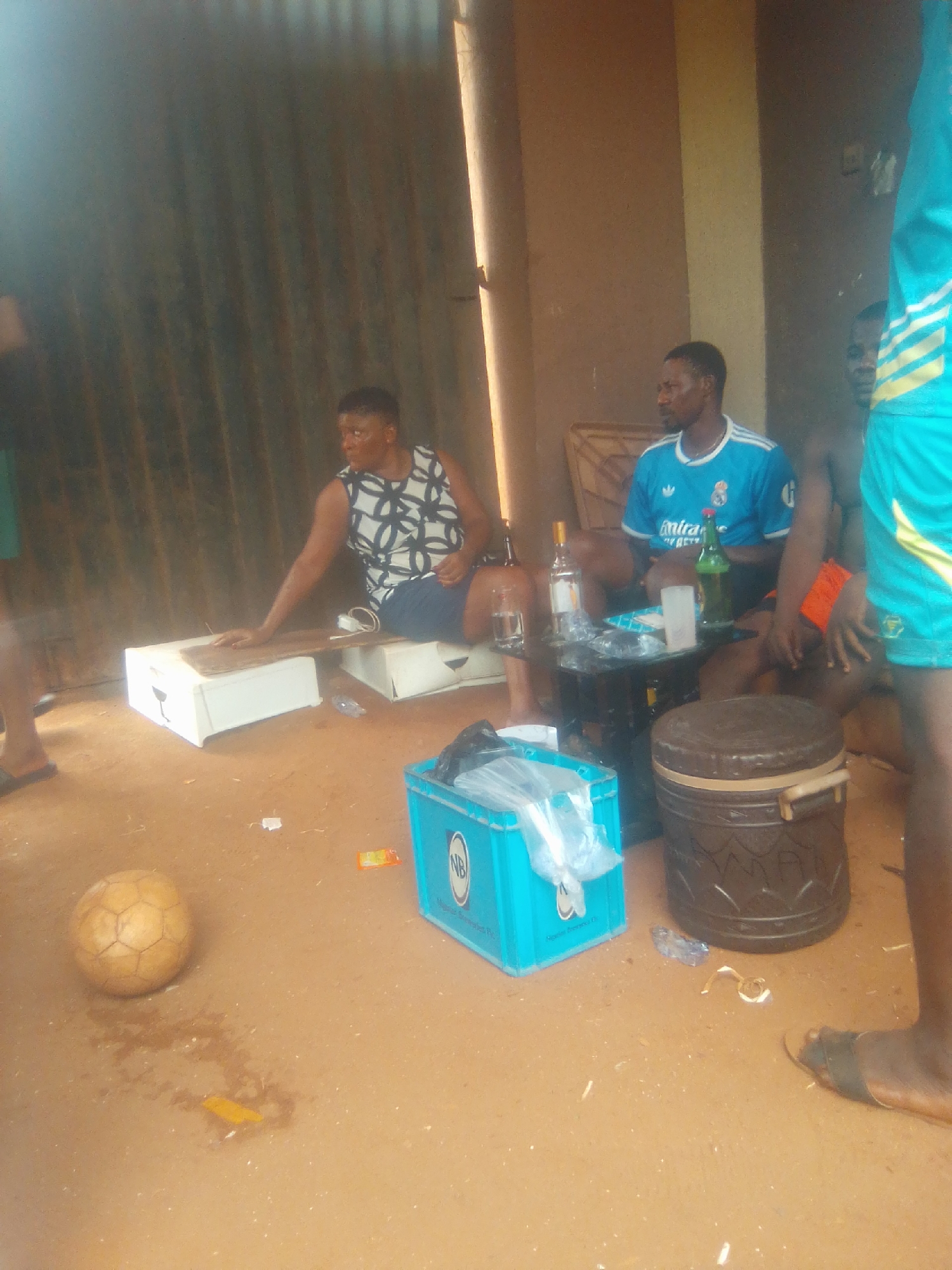 Nsugpe, Nigeria. A corrugated metal structure encloses a gathering. An adult female in a patterned tank top and dark shorts sits on two white plastic boxes. A soccer ball rests on the ground. A blue plastic beer crate with clear plastic bags sits near a brown cylindrical container. A small black table holds bottles, glassware, and a package. An adult male wearing a blue Real Madrid t-shirt is seated at the table. Another adult male is to his right, wearing orange shorts. A fourth adult male is partially visible, wearing a green and yellow outfit. People are sitting, interacting with each other, and objects are scattered around.
