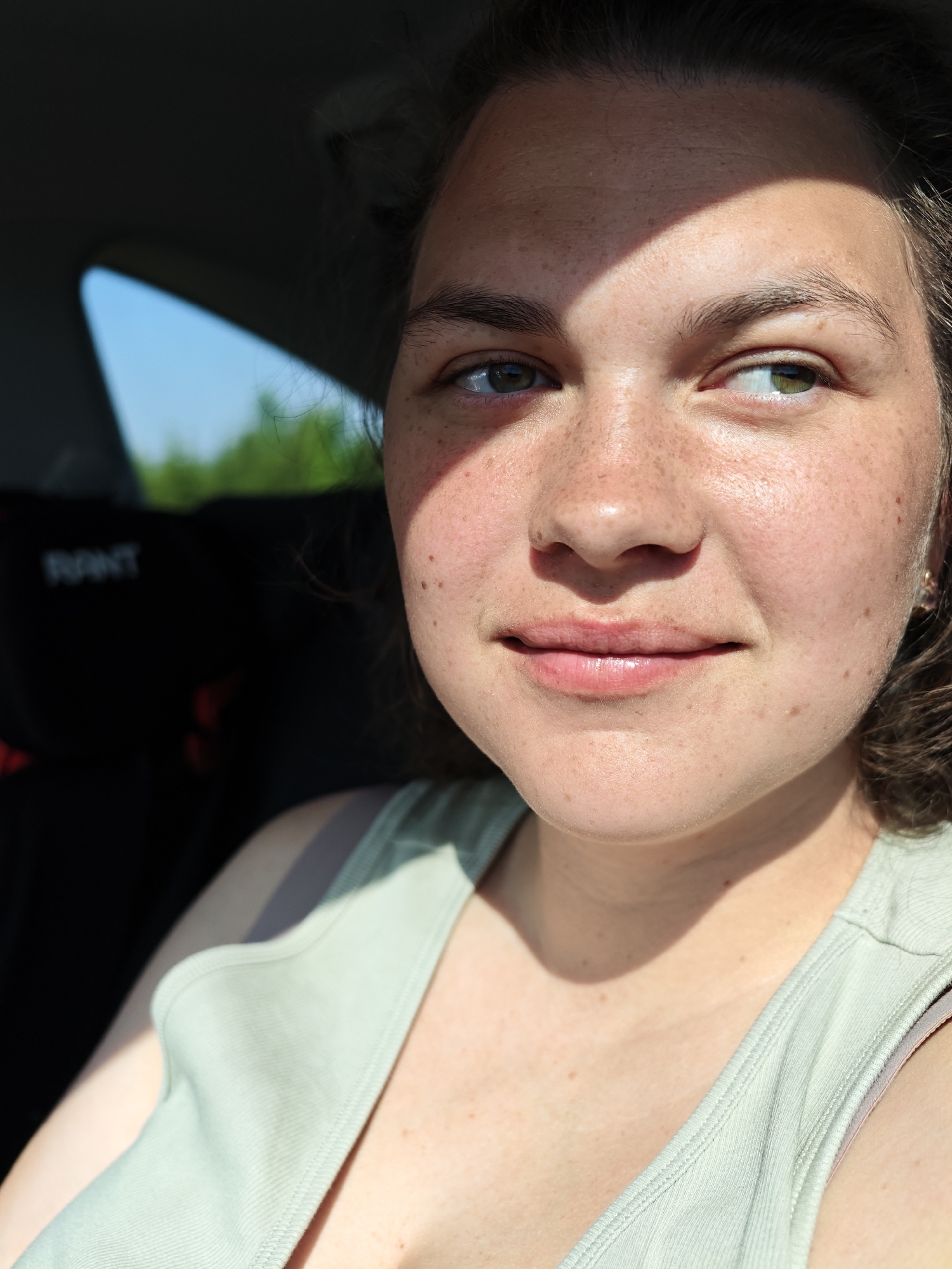 A close-up shot shows a woman with light skin and freckles, partially shadowed on her forehead. She has green eyes and a slight smile. She's wearing a light green tank top. The background is out of focus, but shows the interior of a car, with the suggestion of a green landscape visible through a window. The location of Izh-Boyki, Russia cannot be verified in the image.