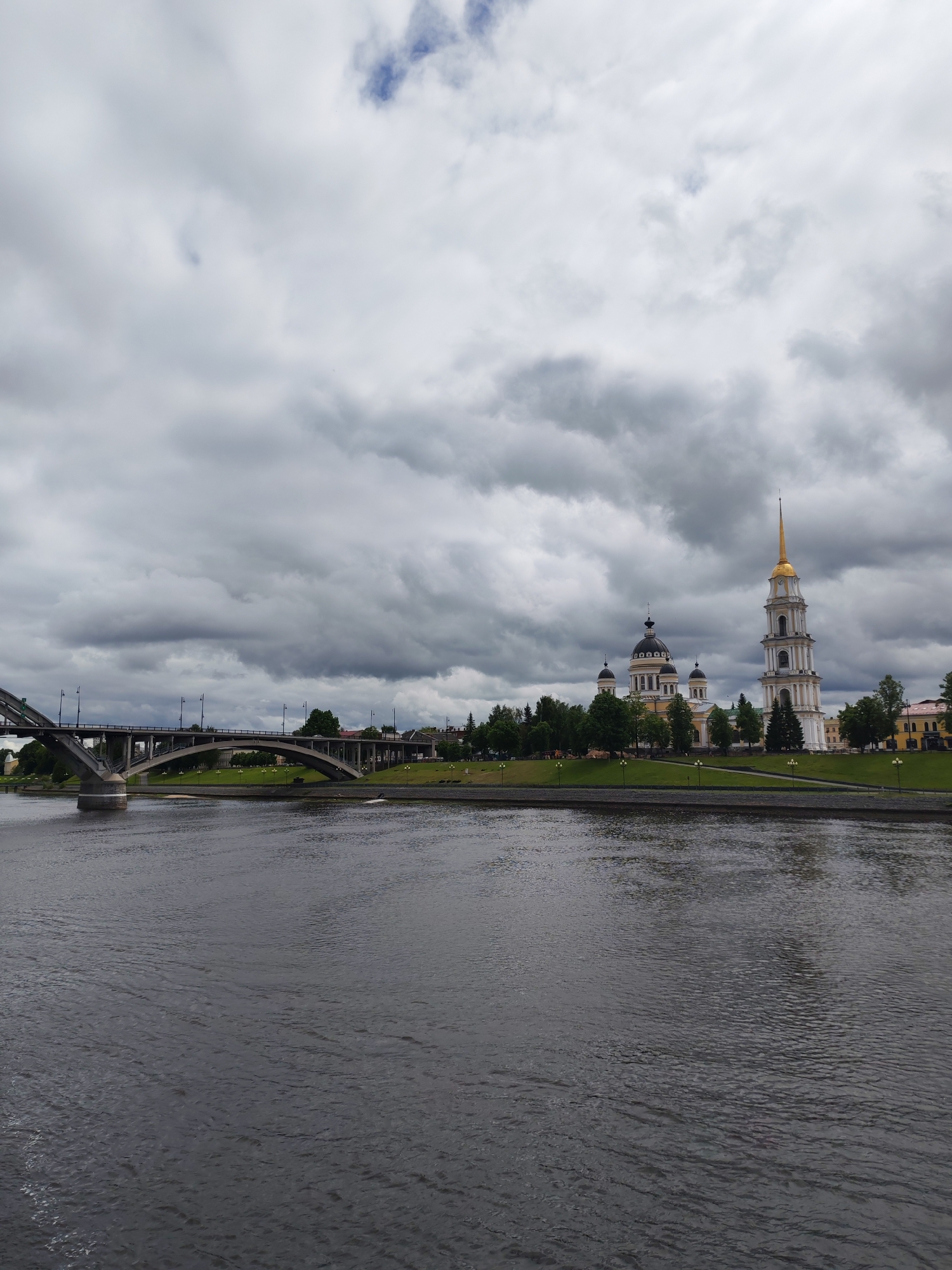 The image captures a cityscape in Rybinsk, Russia, under a heavily clouded sky. A wide river dominates the foreground, its surface reflecting the somber light. To the left, a bridge spans the river, its arched concrete structure leading towards the opposite bank. On the right side of the riverbank, a prominent architectural complex stands, featuring a yellow and white cathedral with black domes and a separate tall, slender bell tower topped with a golden spire. A grassy bank with scattered trees stretches along the river’s edge, interspersed with lampposts. A few other buildings and more trees are visible farther inland. The overall atmosphere is muted, owing to the overcast sky and dark water.