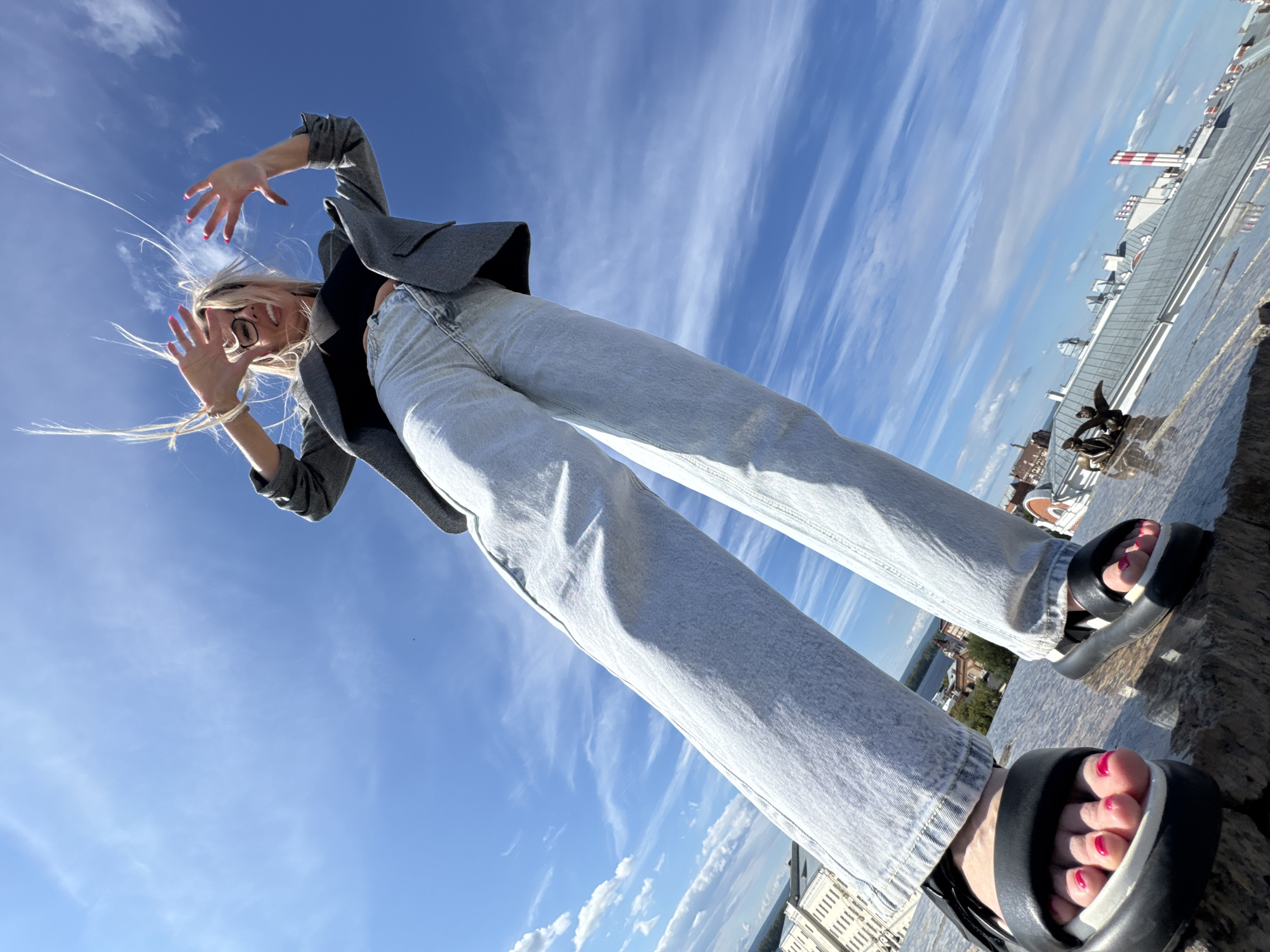 A low-angle shot captures a young woman from the waist down, standing on a stone ledge. She is wearing light-wash wide-leg jeans and black platform sandals with white soles. Her feet are adorned with bright pink nail polish. She is looking up, with her arms outstretched towards the camera, her hair blowing in the wind. The background is a bright blue sky filled with wispy white clouds. In the distance, a cityscape with buildings and a river can be seen. The sun appears to be shining, suggesting it is daytime. The woman's pose and expression convey a sense of playfulness and freedom.
