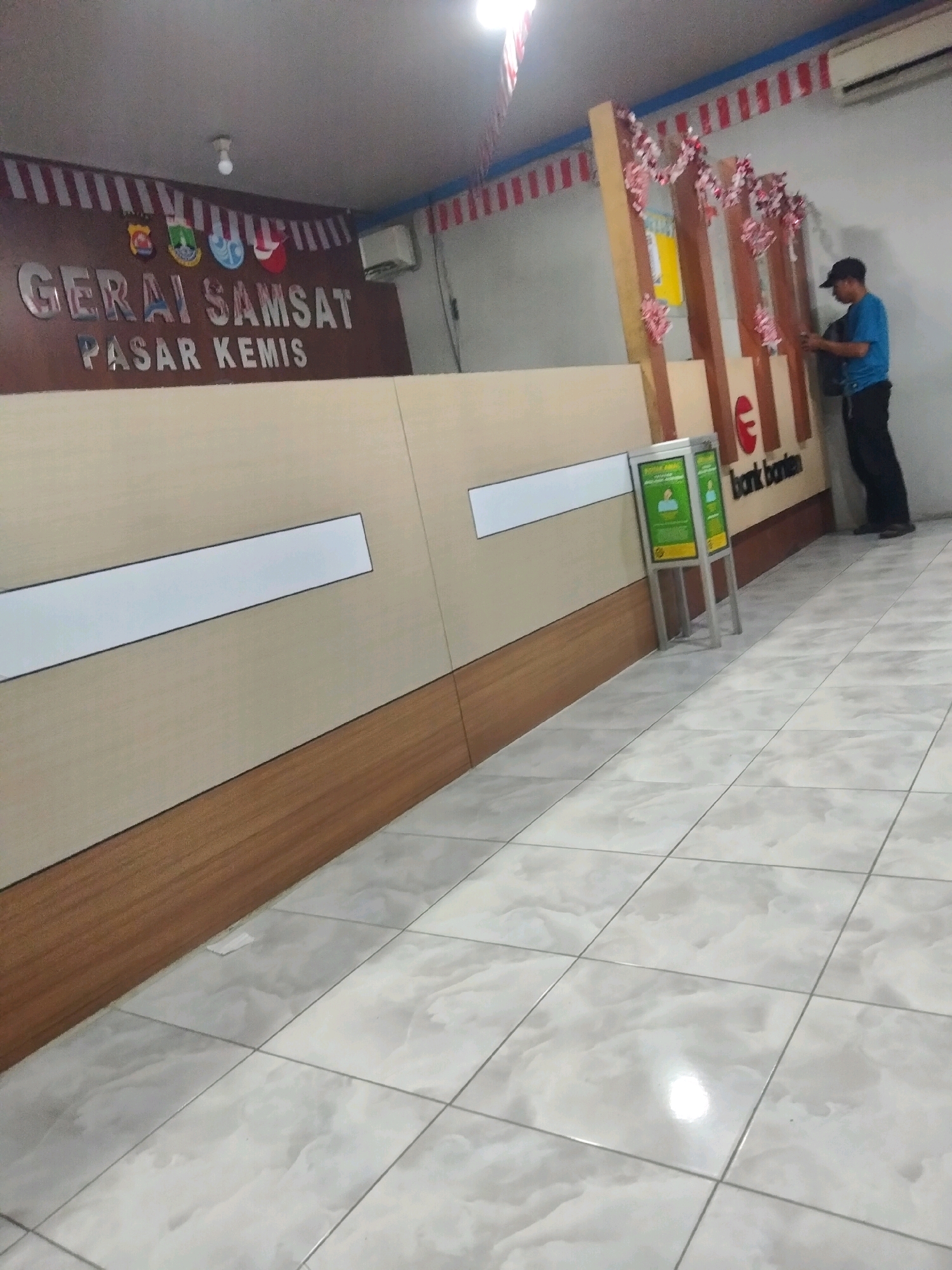 A man in a blue shirt and black pants, wearing a black cap, stands at a counter in a well-lit indoor space. He appears to be interacting with something on the counter, possibly a computer or documents. The counter is made of light-colored wood and has a brown trim at the bottom. Behind the counter, a sign reads "GERAI SAM... PASAR KEMIS" with some emblems above it. Further along the counter, a logo with a red "C" and text that appears to say "bank banten" is visible. To the left of the man, there is a display stand with green panels and images of people. The floor is tiled with a light gray marble pattern. Red and white striped decorations are visible along the top of the wall, suggesting a festive or official occasion. An air conditioning unit is mounted high on the wall. The overall impression is that of an administrative office or a service center in Sukaasih, Indonesia.
