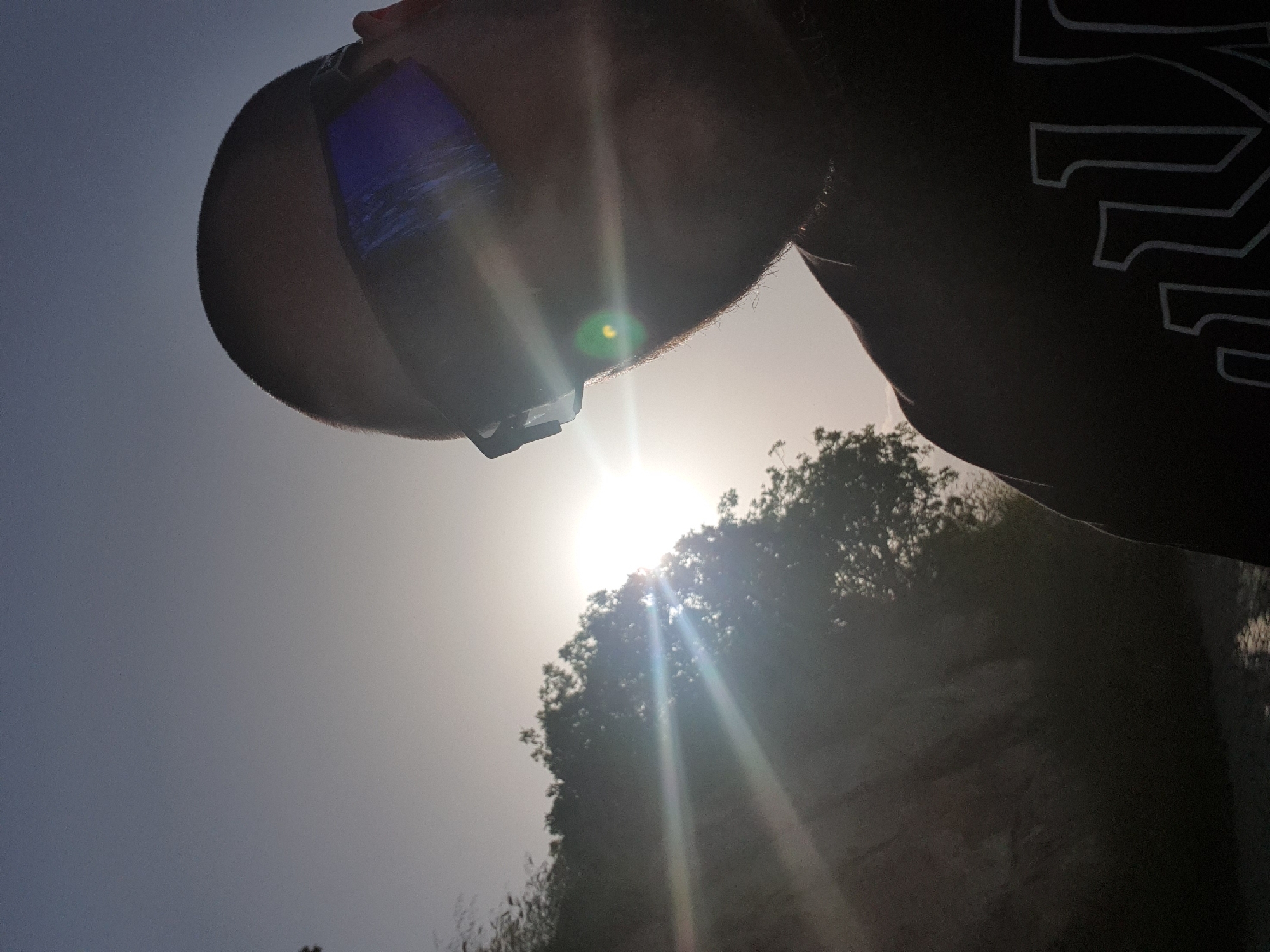 A low-angle shot depicts a person in Plovdiv, Bulgaria. The subject's head fills the upper left quadrant, partially obscuring a bright, sunlit sky and the tops of trees. The person is wearing a smartphone attached to their forehead. The subject is also wearing a black T-shirt. The foreground consists of trees and the ground. The setting appears to be outdoors during a sunny day.