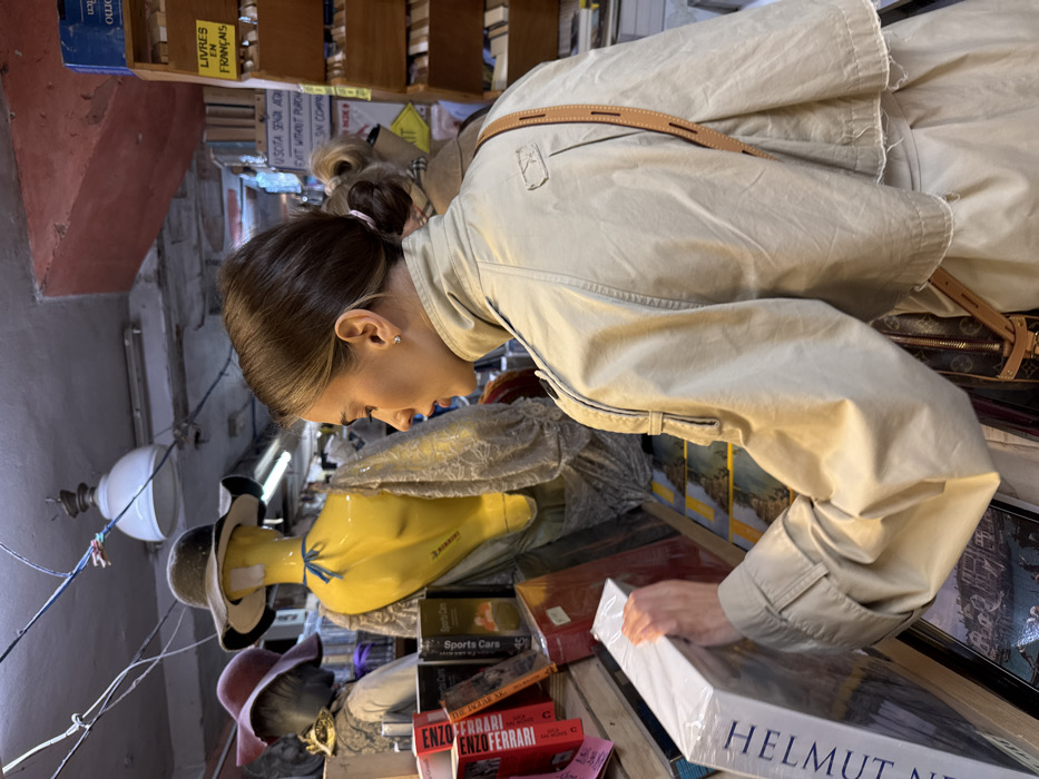 The scene depicts the interior of Libreria Acqua Alta in Venice, Italy. A young adult female is positioned in profile, facing left, examining a large, shrink-wrapped white book, partially labeled "HELMUT NEWTON," which she holds with both hands. Her brown hair is pulled back into a low bun, secured with a light pink scrunchie, and she wears a small stud earring in her visible right ear. She is dressed in a beige trench coat featuring shoulder epaulets and a short, cape-like overlay. A brown leather-strap bag, with a visible monogrammed pattern, hangs across her body from her right shoulder.
To her left and slightly behind her, two mannequins are visible: a headless yellow torso mannequin wearing a dark fedora and a lace shawl, and a partially obscured grey mannequin head wearing a wide-brimmed maroon hat. Bookshelves densely packed with various books, including titles like "ENZO FERRARI" and "Sports Cars," fill the background. Prominent yellow signs are visible, one reading "USCITA SENZA ACQUISTO / EXIT WITHOUT PURCHASE" and another, further back, stating "LIVRES EN FRANÇAIS." The low ceiling shows exposed electrical wiring and features a white industrial pendant light fixture and a fluorescent strip light. A second, partially obscured individual is visible in the distant background.