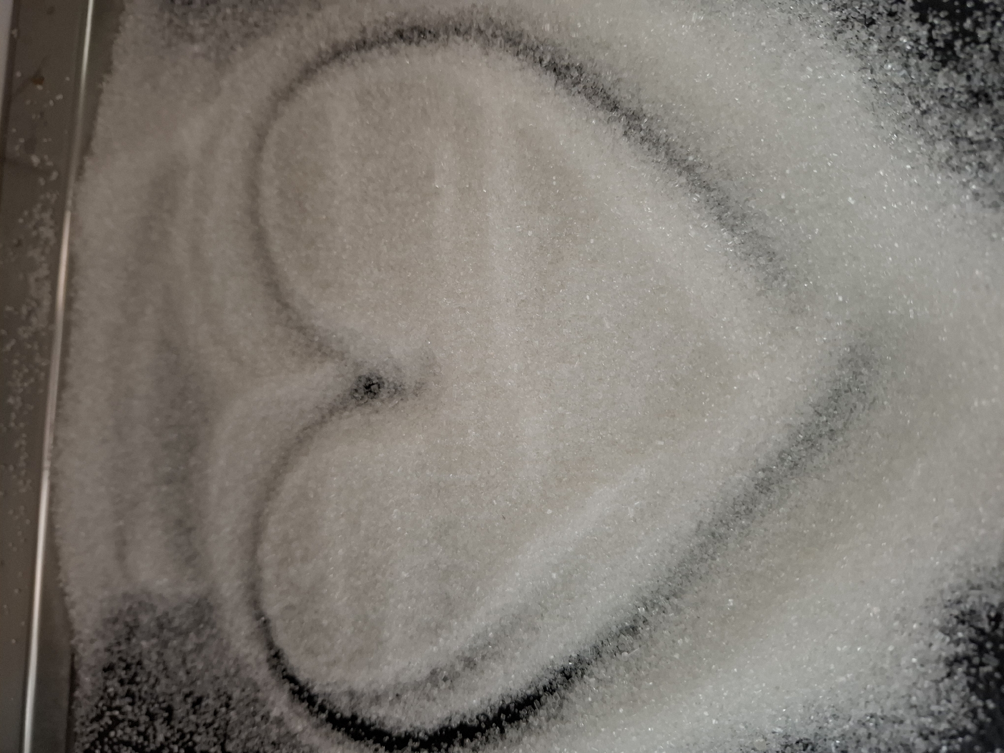 A scattering of white granulated sugar is arranged on a dark surface, likely a tray. A heart shape has been drawn or formed within the sugar, revealing the dark surface beneath. The background is blurred. There's no discernible information to indicate the location in the city of Novi Sad, Serbia. There are no people or interactions visible in this close-up image.