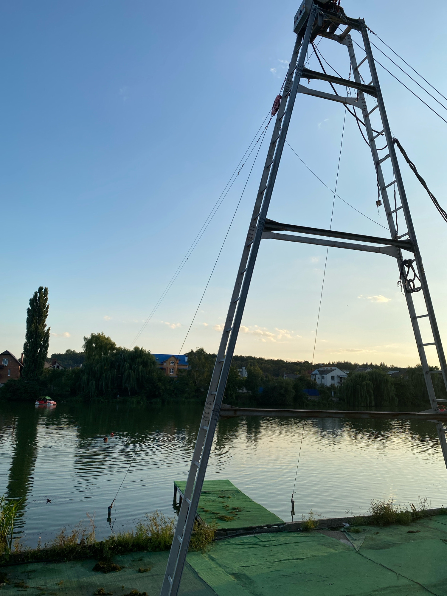 The image depicts a wake park facility located on a body of water, likely a lake or river, in Vinnytsia, Ukraine. A tall, metal A-frame structure, characteristic of a wakeboarding cable system, dominates the foreground and mid-ground on the right side of the frame. The structure is equipped with cables, pulleys, and a ladder for access.
On the water, two individuals are visible, appearing to be wakeboarding or swimming. One person is further out, and another is closer to the shore. In the background, a residential area with houses and lush green trees lines the opposite bank. A unique, car-shaped boat is also present on the water near the left bank, adding a whimsical touch to the scene. The sky is clear with a few scattered clouds, indicating fair weather, and the sunlight suggests it is either late afternoon or early evening. The overall environment is recreational, with elements of nature and man-made structures integrated for water sports activities.