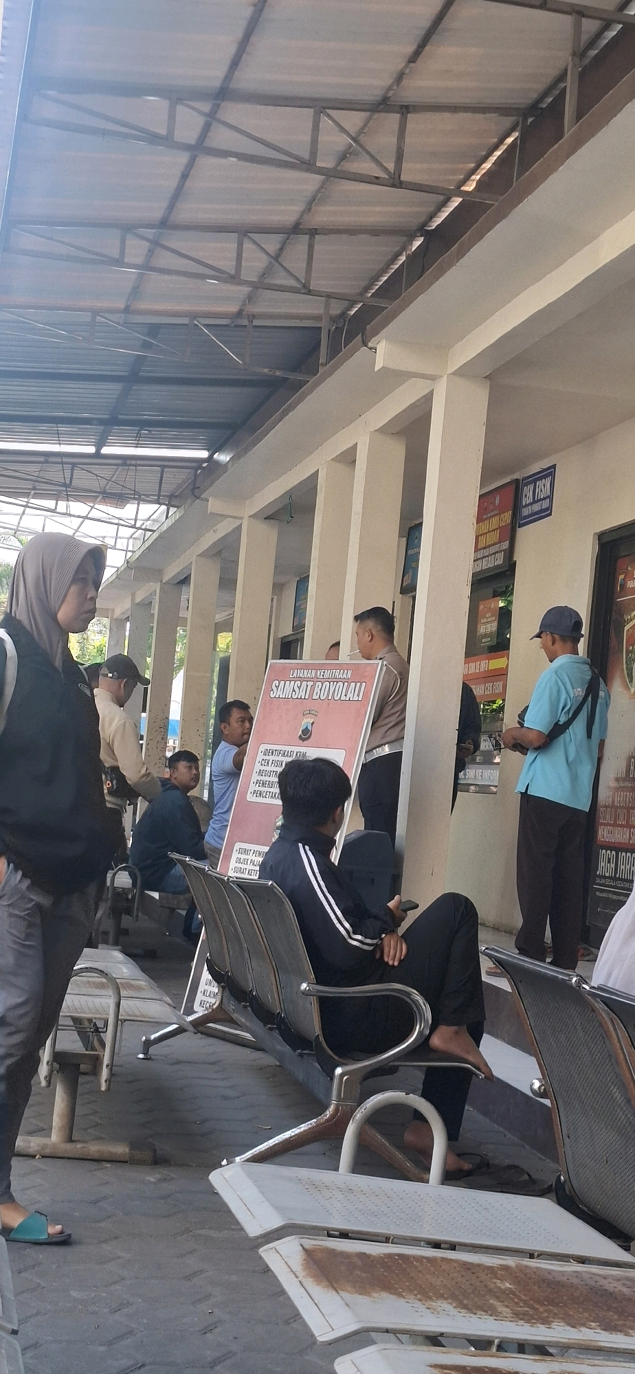 The image depicts an outdoor waiting area, possibly at a government service center, in Gumulan, Indonesia. A group of people are seen queuing or waiting to be served. In the foreground, a woman wearing a grey hijab and dark clothing is looking to her left. Behind her, a man in a light-colored shirt and dark pants is also visible.
Further back, a sign with red text reads "LAYANAN KEMITRAAN SAMSAT BOYOLALI," indicating that this is a vehicle tax service center (SAMSAT) in Boyolali, a town near Gumulan. Several individuals are standing near the sign and the service counter. One person in a light blue shirt appears to be interacting with the counter. Another man in a dark jacket is seated on a metal bench, looking to his right.
The environment consists of a covered walkway with a metal roof structure supported by columns. The floor is paved with dark tiles. Several rows of metal benches are arranged for waiting individuals. In the background, there are more structures and signs, suggesting a government administrative or service building. The overall scene conveys a sense of a busy service center during daytime hours.
