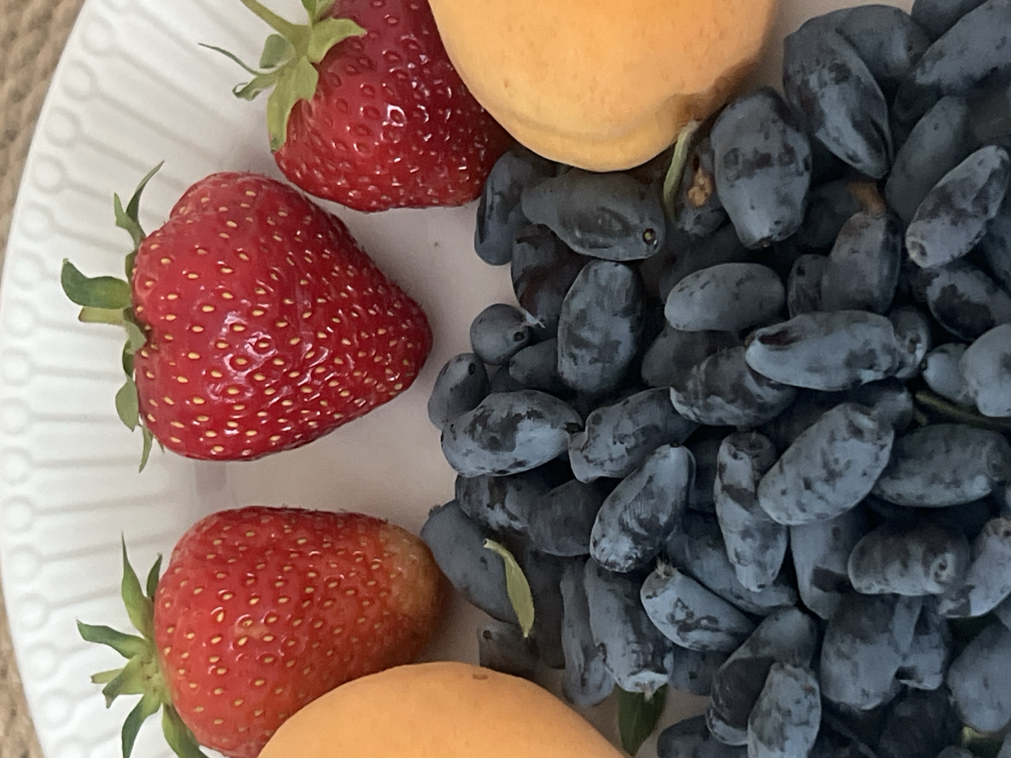 The image displays a close-up view of a white plate filled with fresh fruits. Three red strawberries are prominently positioned at the top, with their green stems still attached. Below the strawberries, a large pile of dark blue, elongated berries fills the majority of the plate. Two orange-hued fruits, possibly apricots or peaches, are partially visible on the left and right edges of the frame. The fruits are arranged in a still life composition, suggesting they are intended for consumption or display. The background is a plain, textured surface, possibly a table or tablecloth. The lighting is soft and diffused, highlighting the textures and colors of the fruits. There are no people or animals present in the image. The scene depicts a simple arrangement of healthy food items. There is no indication of the city of Votkinsk, Russia, or any other specific location in this image.