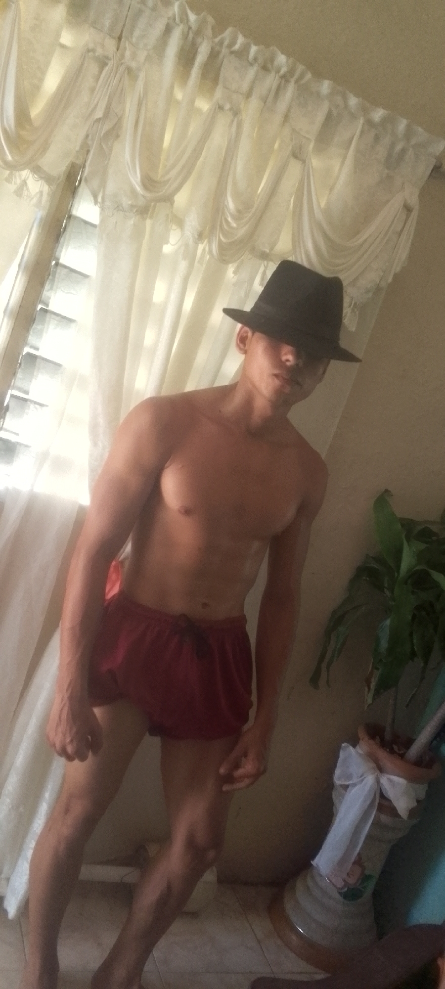 A shirtless, fair-skinned male stands in a room with a black fedora covering his forehead and eyes. He wears short, maroon athletic shorts. Behind him are white curtains, a window, and a large potted plant with a white bow tied around the planter. The setting appears to be indoors. The image does not provide any conclusive evidence of its location in Juan Griego, Venezuela.