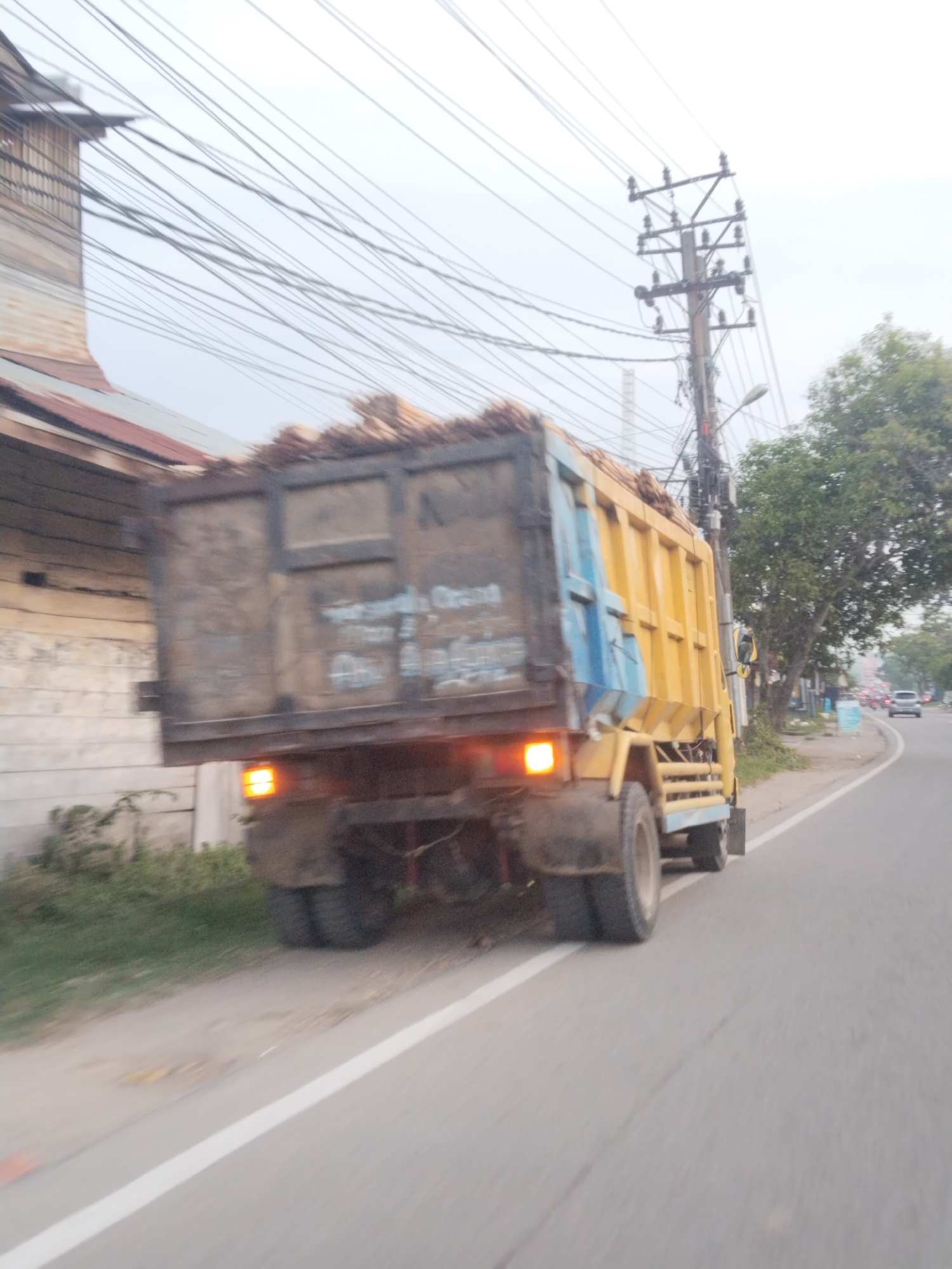 A yellow dump truck is traveling on a paved road in Lhokseumawe, Indonesia. The truck is loaded with palm oil fruit bunches and is partially obscured by motion blur. The truck has a dark-colored cargo bed with blue accents. The road is lined with a grassy verge and some vegetation. On the left side of the road, there is a weathered building with a corrugated metal roof. Overhead, a tangle of power lines crisscrosses the sky, supported by wooden utility poles. In the background, trees and other vehicles are visible, and the sky is overcast. The image captures a scene of everyday transportation and infrastructure in an Indonesian city.
