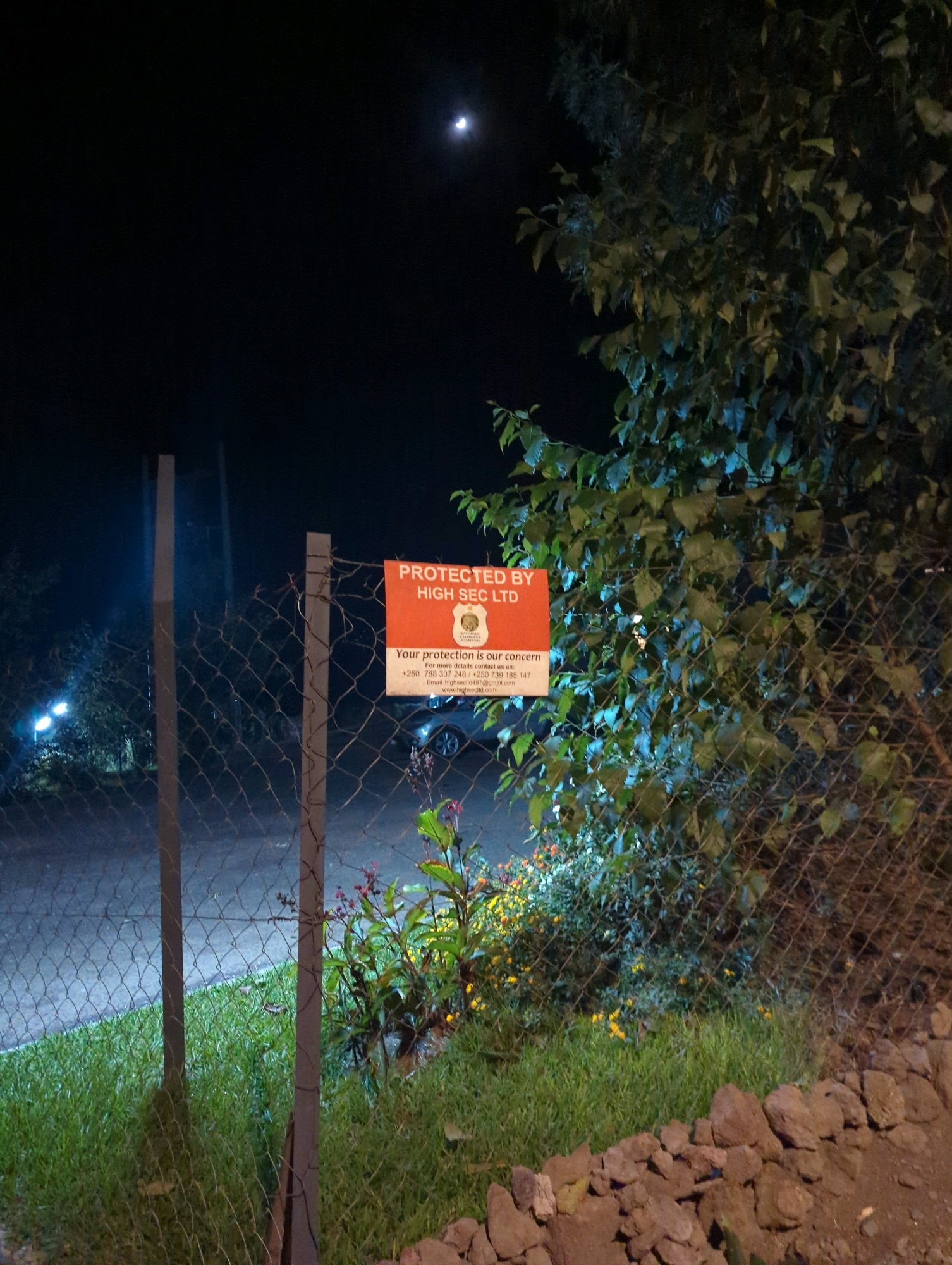 This is a nighttime photograph taken in the Musanze District of Rwanda. The scene is dimly lit, with a crescent moon visible in the dark sky. A chain-link fence runs across the foreground, with two metal posts supporting it. Attached to the fence is a rectangular orange sign that reads "PROTECTED BY HIGH SEC LTD." Below this text, there is a crest and smaller text providing contact information, including a phone number and email address. Behind the fence, there is a patch of green grass and some flowering plants with yellow and red blooms. A large tree with dark green leaves is situated to the right of the frame, partially obscuring the view of the background. In the distance, faint lights from what appears to be a road or street are visible, along with the silhouette of a parked vehicle. The overall environment suggests an outdoor area, possibly a residential or commercial property, secured by a security service.