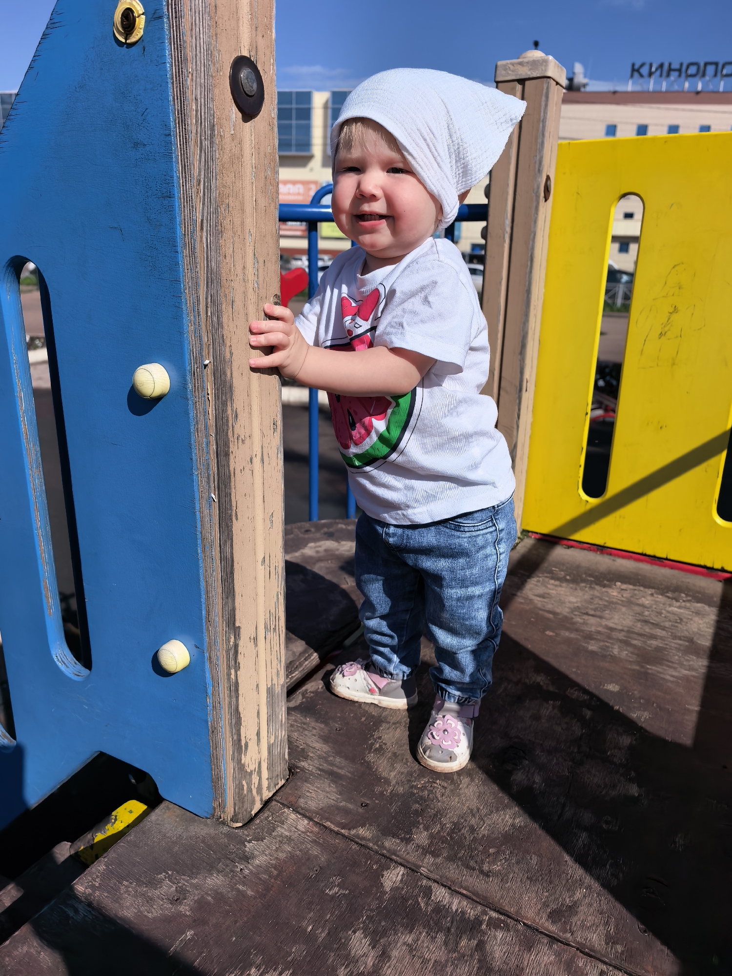 An infant stands on a wooden platform of a playground structure, against a backdrop of a building with Cyrillic lettering that appears to say "КИНОПО..." likely indicating a cinema. The child is wearing a white bandana on their head, a white t-shirt with a watermelon graphic, and blue jeans. The playground structure is composed of blue and yellow panels and wooden beams. The sun is shining, casting shadows on the wooden surface. The sky is blue with some clouds. The scene is outdoors, likely in a public area of Sterlitamak, Russia.