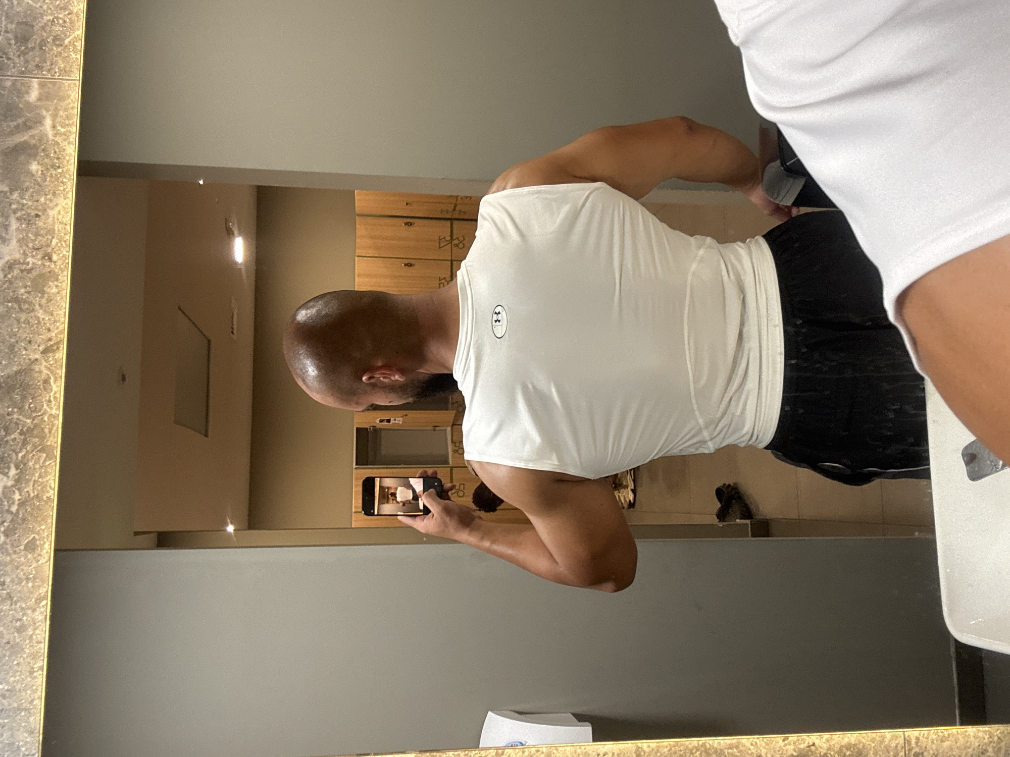 A bald, muscular man is taking a selfie in a gym locker room. He is wearing a white tank top and black shorts. The locker room has wooden lockers and a tiled floor. There is a sink in the foreground and a mirror reflecting the man. The scene appears to be in Sofia, Bulgaria.