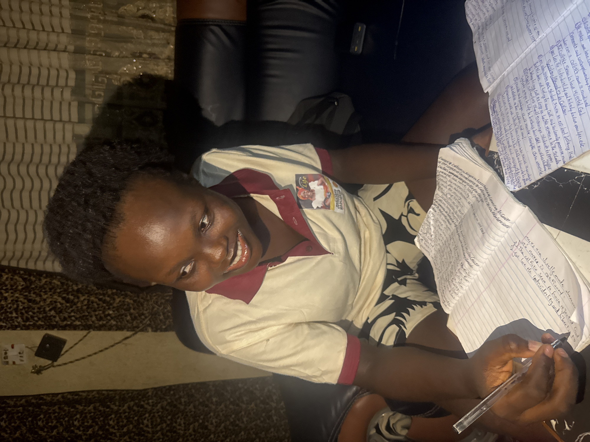A young woman is seated, writing on lined paper with a clear pen. She is smiling and looking up. She is wearing a light-colored, short-sleeved shirt with maroon trim and a small badge pinned to it. The background includes a black leather sofa, a striped rug, and additional papers. Text is visible on the papers. There are power adapter and cord on the rug. Based on the badge on the shirt and reference to Nigeria, it is possible the location is Onitsha, Nigeria, but that cannot be confirmed from the image alone.