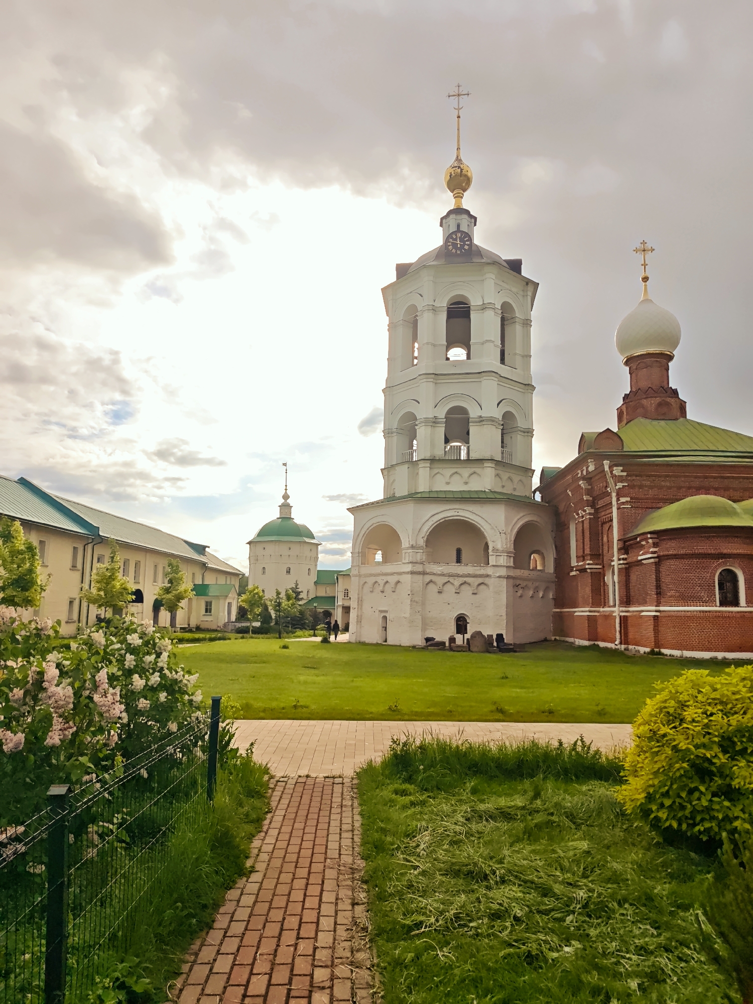 The image captures a serene courtyard scene within a monastery complex, likely located in the city of Луговой, Russia. A brick-paved pathway leads the viewer's eye toward the architecture. The courtyard features green lawns.
On the right stands a brick-red building with green roof and white dome topped with a cross, characteristic of Russian Orthodox architecture. Adjacent to it is a white, multi-tiered bell tower, complete with arched openings and a clock face under a golden spire.
To the left, a lower building with a light-colored facade and a green roof extends into the background. The sky is overcast with clouds. The presence of lilac bushes and other greenery adds life to the environment.