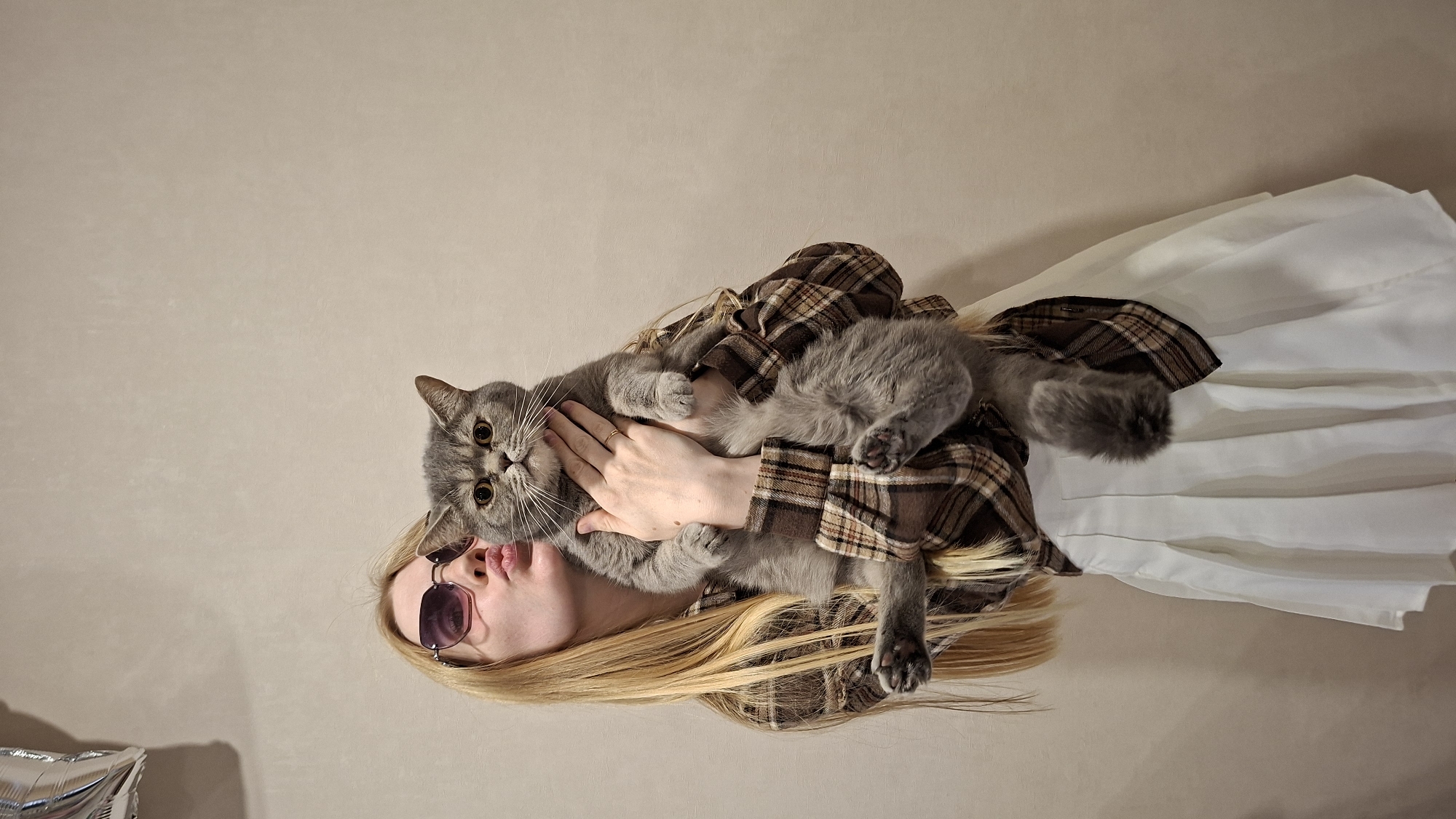 A young woman with long blonde hair and sunglasses is lying down, holding a grey cat. The woman is wearing a brown and white plaid shirt and a white skirt. The cat is lying on top of the woman, wrapped around her torso. The woman's hair is spread out around her head. The background is a plain, light-colored wall. There is a silver balloon partially visible in the lower left corner. The image is taken from above, looking down at the woman and cat. The lighting suggests it is daytime. The scene appears to be indoors, likely in a home. The woman has her lips pursed, and the cat is looking directly at the camera with its eyes wide open. The overall mood is playful and intimate.