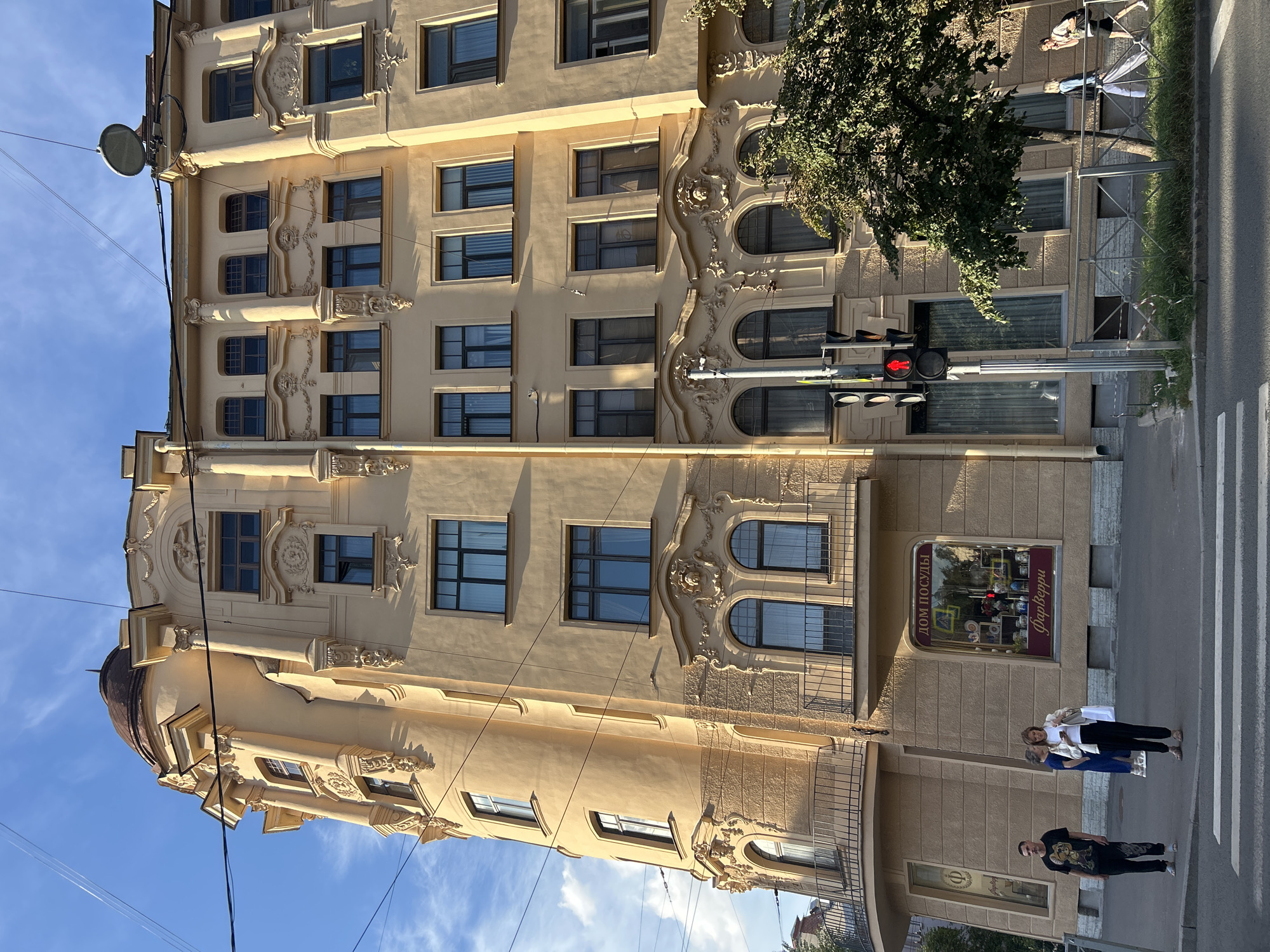 The image depicts a street scene in Saint Petersburg, Russia, on a sunny day with a few clouds. The primary focus is a large, ornate, yellow building with several stories, featuring a curved facade, decorative architectural elements, and numerous windows. The building has a domed roof on its left side and a satellite dish on its right.
In front of the building, there is a sidewalk and a street with a pedestrian crossing. Several people are visible:
* Two men are standing on the sidewalk, one in a black t-shirt with a graphic and patterned pants, the other in a black t-shirt and patterned pants.
* A woman in a blue dress and white jacket, carrying a white tote bag, is standing on the sidewalk and looking at her phone.
* Two other individuals are walking along the sidewalk on the right side of the image, near a tree.
A traffic light with a red arrow illuminated is visible on a pole in front of the building. A tree with green foliage is located to the right of the traffic light. The building has a shop window with signage that reads "ДОМ ПОСУДЫ" (House of Tableware) and "Фарфоровые" (Porcelain). Overhead power lines are present in the sky. The overall scene is a typical urban environment with pedestrian activity and architectural interest.