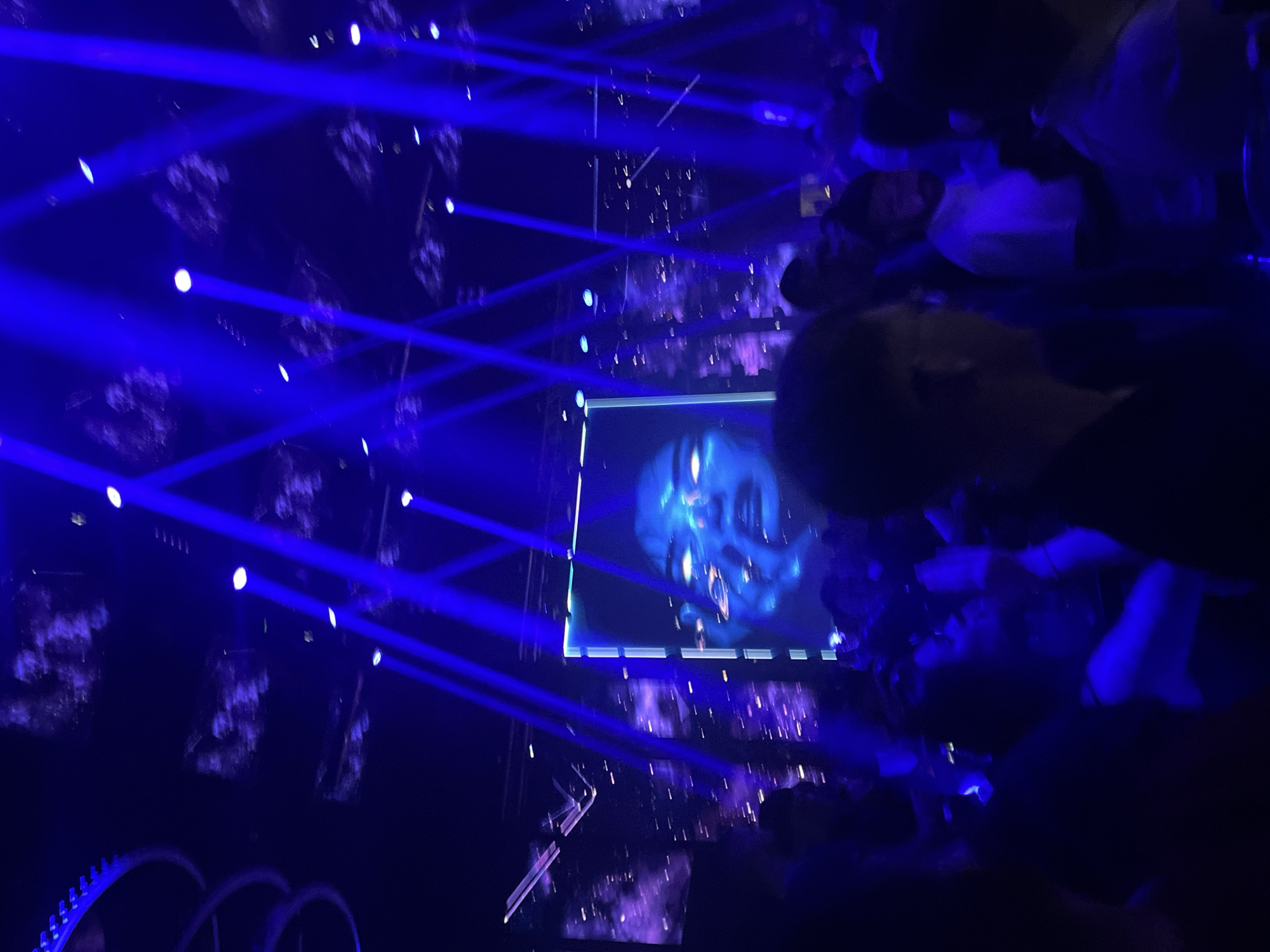 The image shows a nighttime scene, possibly a concert or performance, likely indoors given the lighting setup. Strong blue laser beams cut through the air. A screen displays a face-like image with multiple eyes, bathed in blue light. The edges of the screen are illuminated. People are present, mainly in the background and towards the right, their features obscured by the low light and angle. Structures resembling decorative elements are visible at the bottom of the image. Given the context, it is reasonable to say this happened in Plovdiv, Bulgaria.