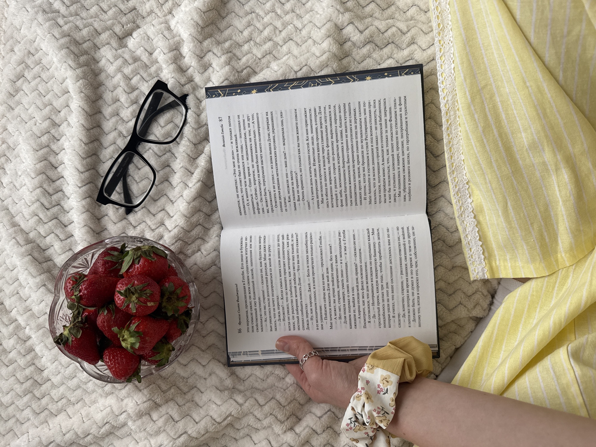 A book with pages open, a bowl of strawberries, eyeglasses, and a scrunchie lay on a textured off-white blanket. A hand with a ring holds the book open. To the right, a yellow striped shirt is visible. The book is open to pages 86 and 87, with text in Russian, and the title "The Great Gatsby". The strawberries are bright red with green stems. The eyeglasses have a black frame. The scrunchie has a floral pattern and is yellow.