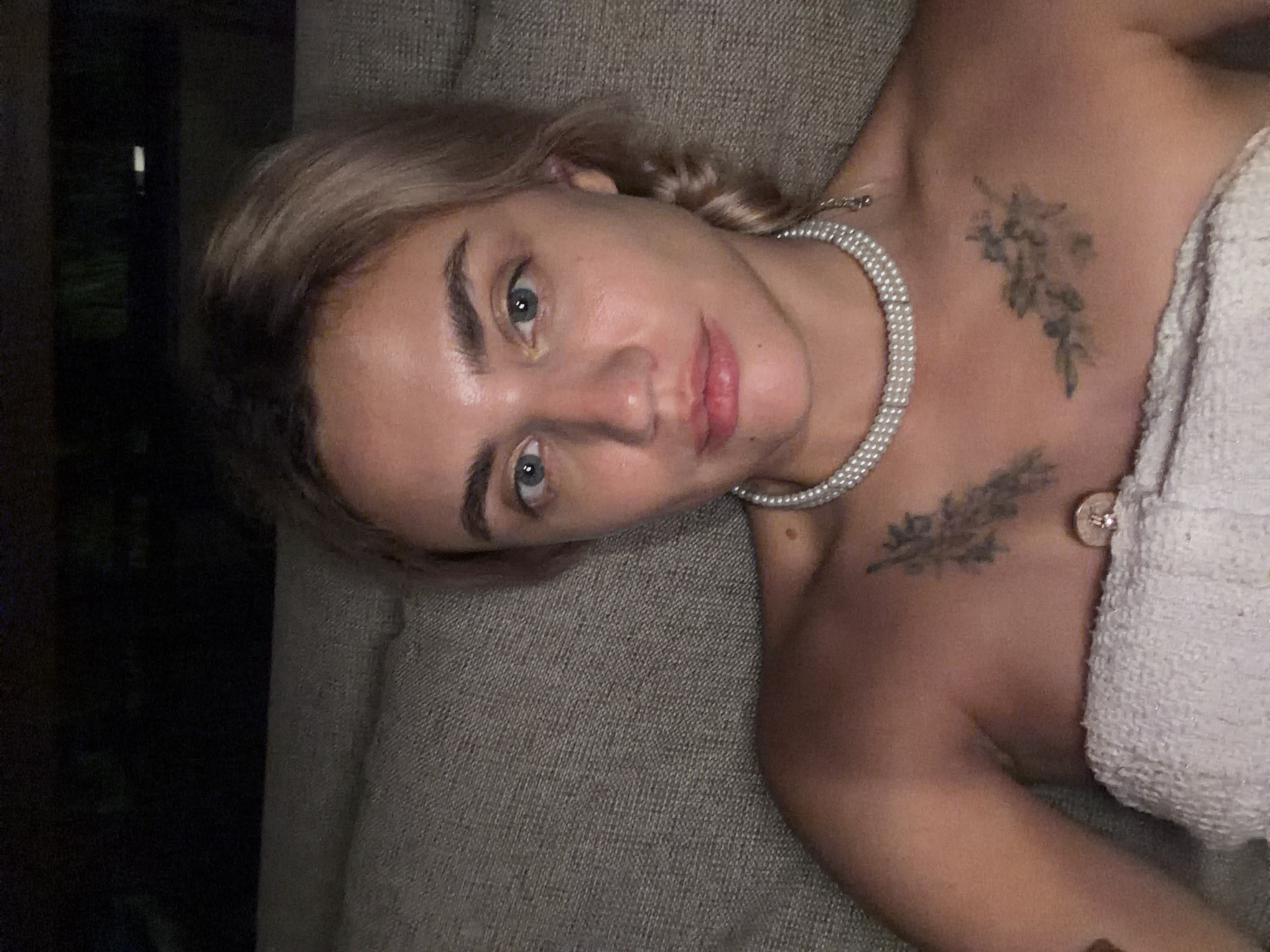 The image shows a woman with light-colored hair and blue eyes. She is wearing a pearl necklace and has two tattoos on her chest, one resembling a feather and the other a cluster of flowers. She is also wrapped in a white towel, and a portion of her shoulder and upper chest are visible. The background is dark and appears to be an indoor setting, possibly a lounge area with a textured surface behind her. The image does not contain any information about the city of Kediri, Indonesia.