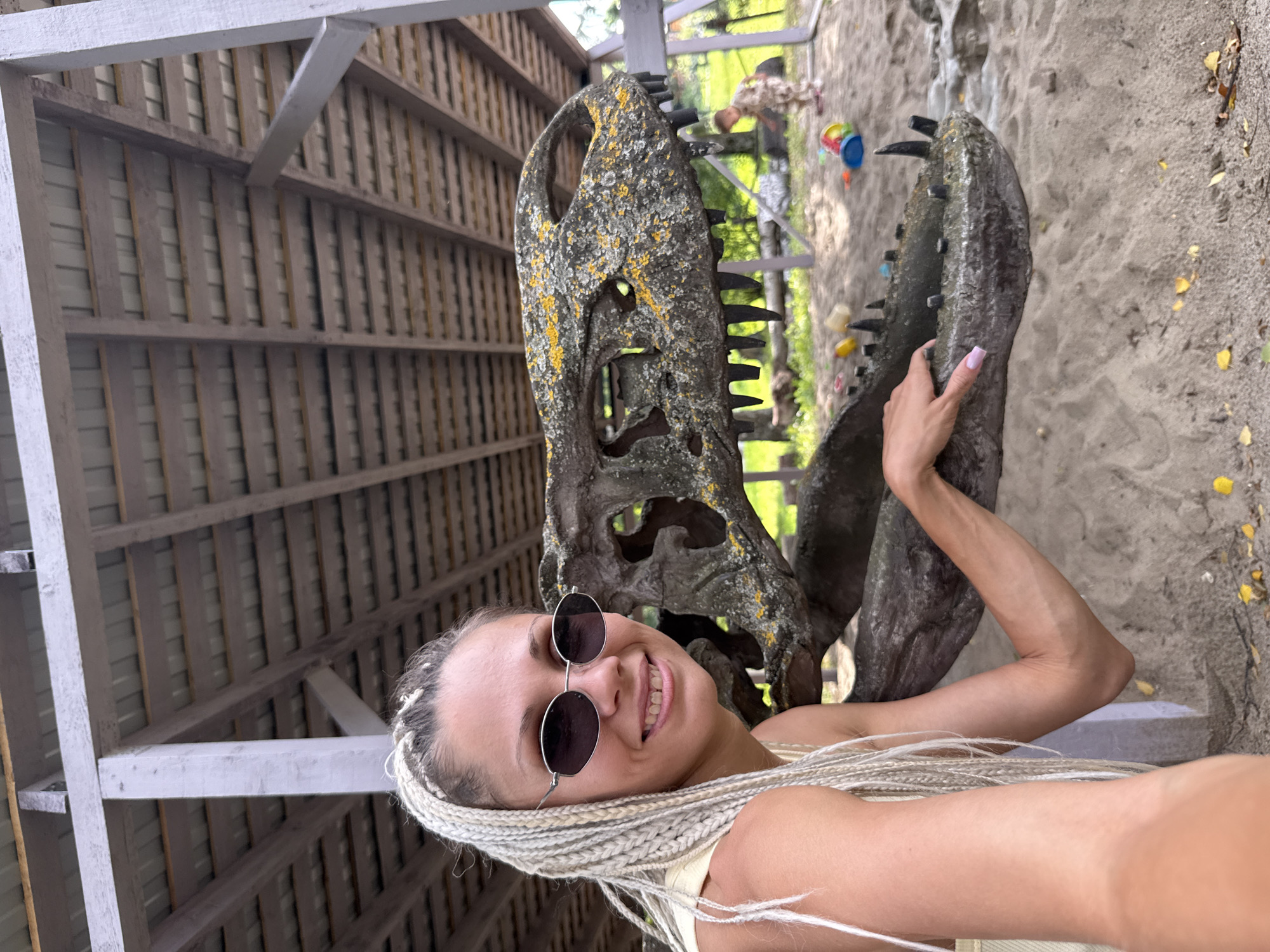 In an outdoor area covered by a wooden roof structure, a woman with blonde braided hair and sunglasses smiles at the camera. She is positioned to the left of a large dinosaur skull, part of which is resting on a sandy surface. Her left arm is extended, with her hand resting on the dinosaur's lower jaw. In the background, a child dressed in camouflage clothing is visible playing in a sandbox with toys. The environment appears to be a park or play area, likely within the city of Zhukovo, Russia, given the context.