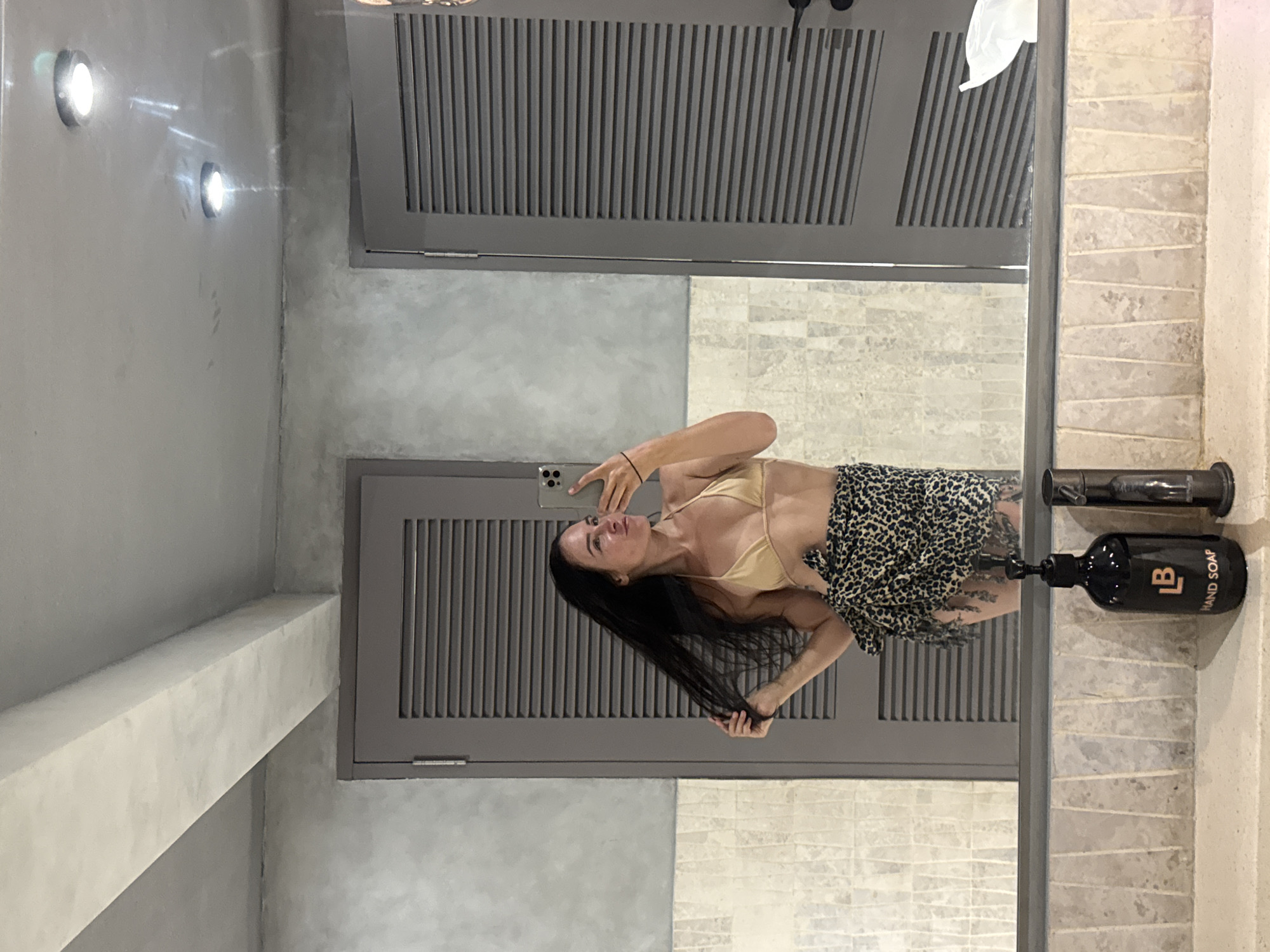 A woman is taking a selfie in a bathroom. She has long dark hair and is wearing a gold bikini top and a leopard-print sarong. She is standing in front of a mirror, and behind her, there are two grey doors with slatted panels. The bathroom walls are a rough, grey concrete finish, and there is a tiled backsplash above the sink. On the counter, there is a black soap dispenser with a gold logo that says "LB HAND SOAP," and a metal faucet. The image appears to be taken in a hotel or resort bathroom in North Kuta, Indonesia.