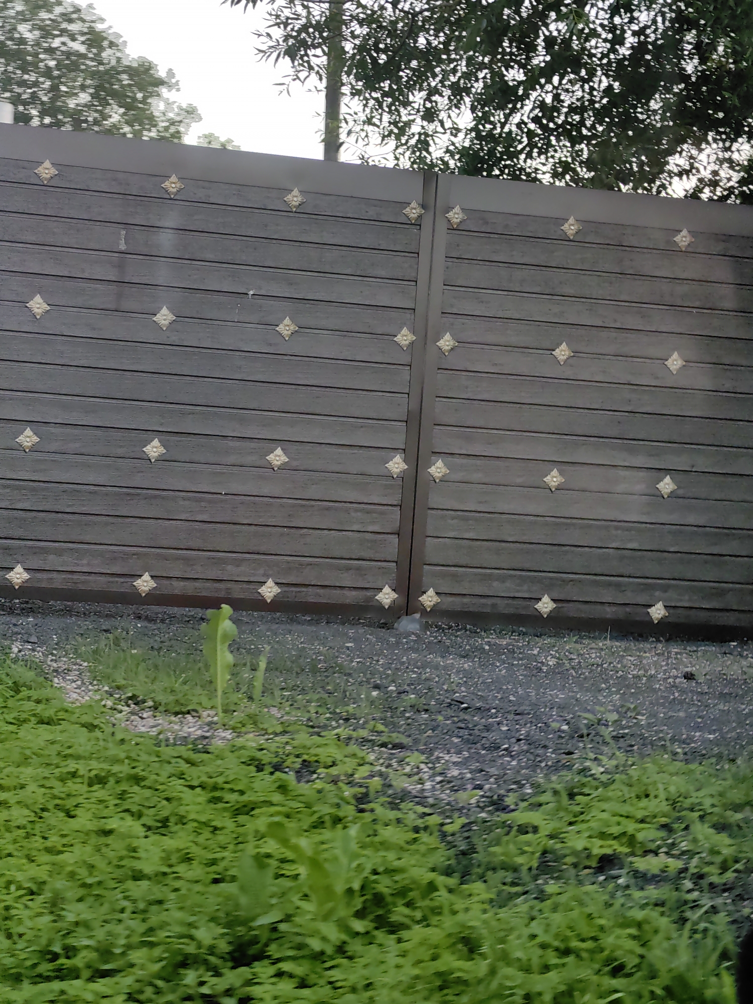 The image shows a section of a dark brown wooden gate with decorative gold rhombus-shaped embellishments. The gate is constructed of horizontal planks. To the left of the gate, there is a patch of green grass. To the right of the gate, there is a gravel area. Overhead, the tops of trees are visible against a bright sky. The scene appears to be outdoors, possibly in a residential or rural setting. No people are present in the image.