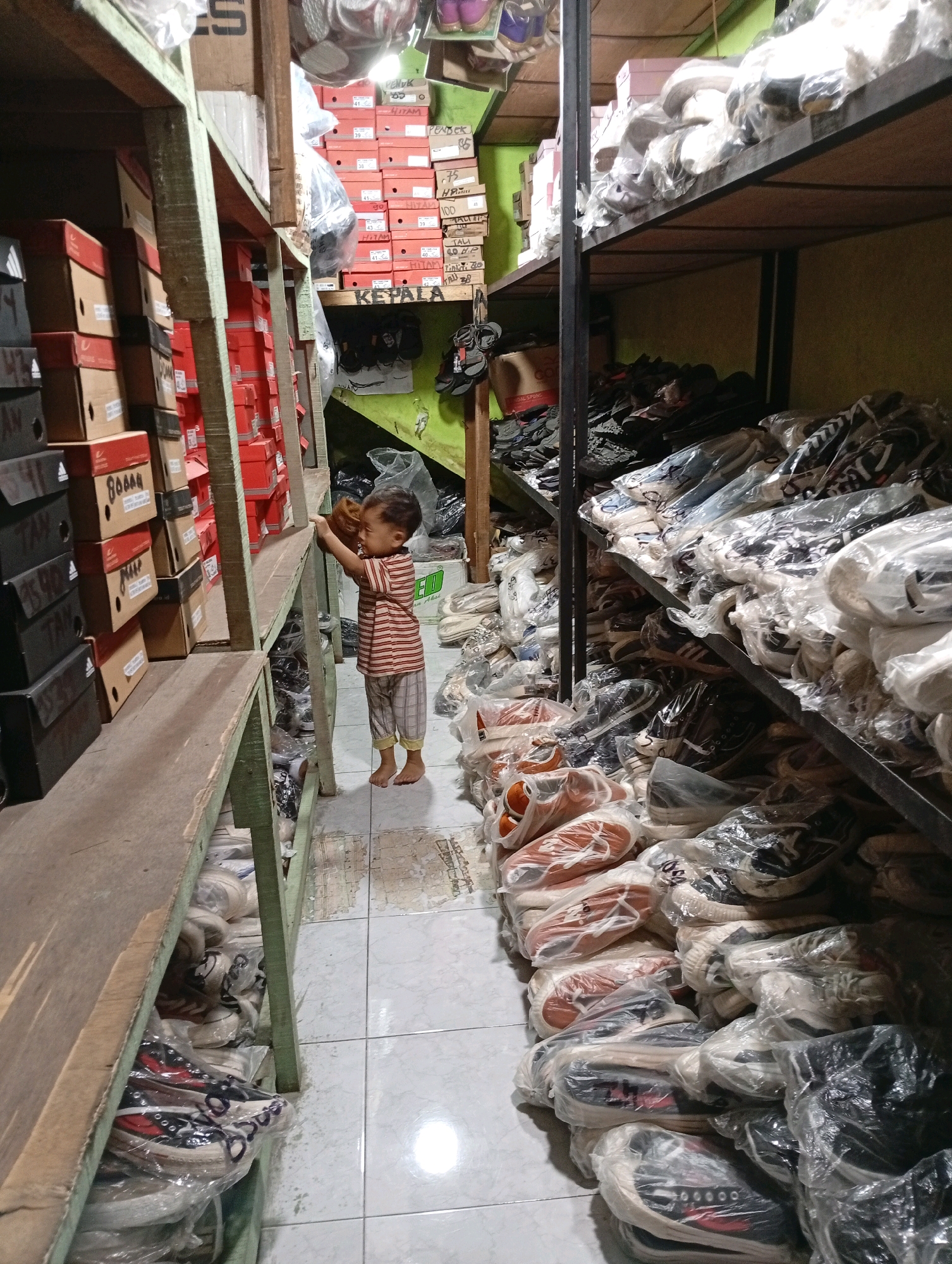 The image depicts the interior of a shoe shop or storage facility in Babakan, Indonesia. The scene is filled with shelves stacked high with boxes of shoes and individual shoes wrapped in plastic. The shelves are made of rough-hewn wood and metal, showing signs of wear and tear. On the left side of the image, stacks of shoe boxes are visible, some with handwritten labels. A young child, wearing a striped t-shirt and plaid pants, stands on the floor between two shelving units, holding a brown object and looking towards the right. The floor is tiled and appears slightly dirty. On the right side, numerous pairs of shoes, primarily sneakers, are arranged on shelves, many still in their plastic packaging. The overall impression is one of a busy, perhaps disorganized, retail or storage space overflowing with merchandise. The lighting is somewhat dim, suggesting an indoor environment with artificial light.
