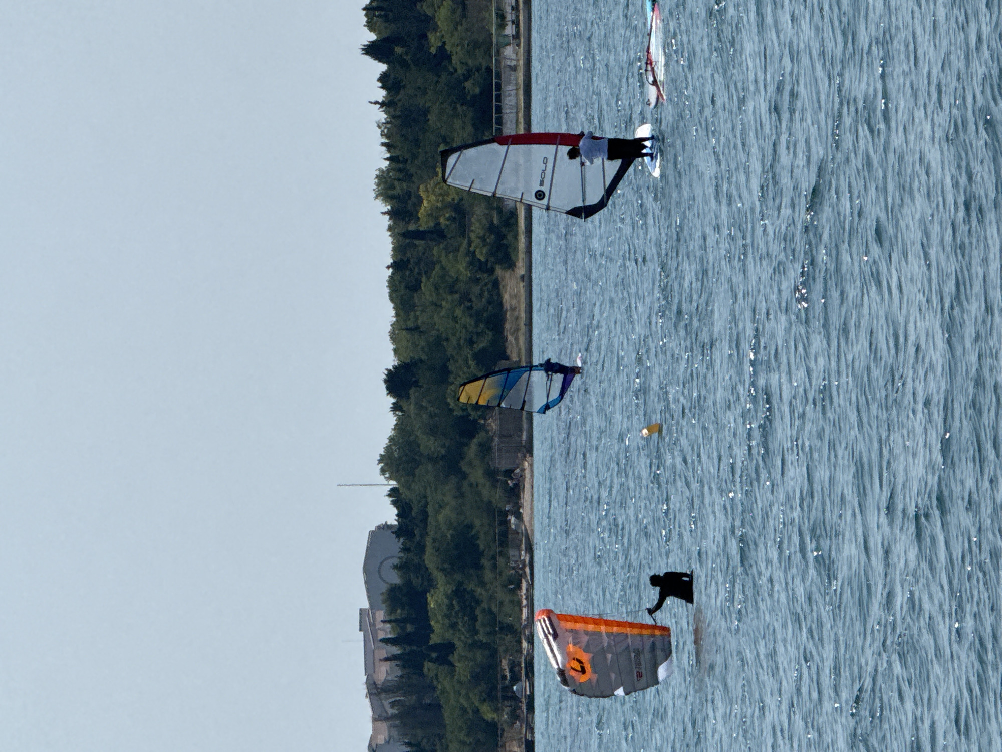 Three individuals are windsurfing on a body of water with a shoreline featuring trees and buildings in the background. The water is choppy, with small waves. One windsurfer, silhouetted in black, is on the left side of the frame, holding an orange and gray sail. In the middle, another windsurfer with a blue and yellow sail is further out on the water. On the right, a third windsurfer, wearing white and black clothing, is on a white sailboard with a red trim. The shoreline displays dense green trees and several buildings, some with curved architectural elements, consistent with the architecture seen in Sevastopol, Ukraine. A buoys floats in the water between the first two windsurfers. The sky is clear and light blue.