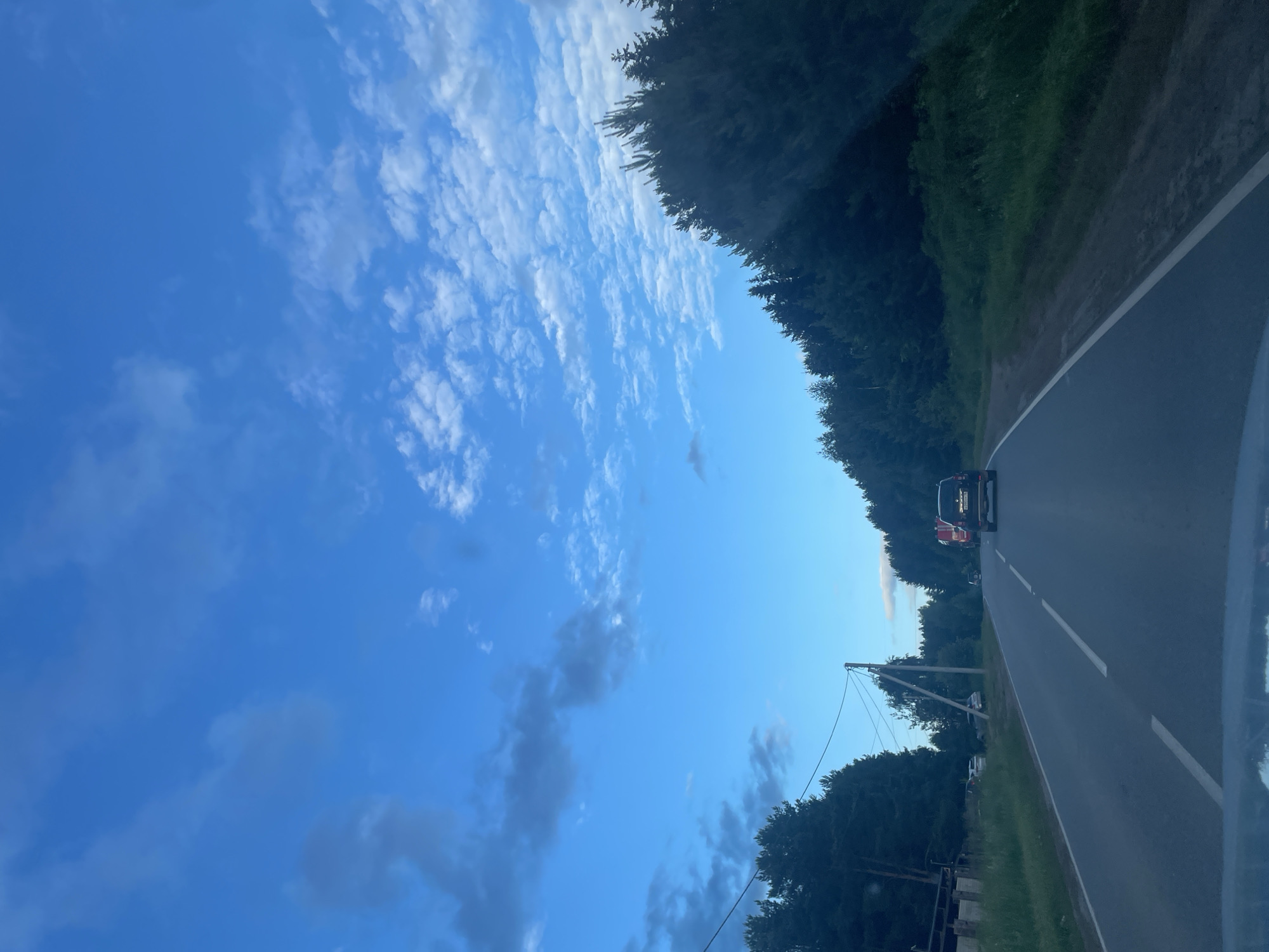 This is a vertical, outdoor, daytime photograph taken from a vehicle traveling on a paved road. The scene depicts a roadway flanked by dense evergreen trees on the left and a grassy verge on the right. A black SUV is visible ahead on the road, with a white license plate. The sky above is a vivid blue, streaked with wispy white clouds. Power lines with poles run along the right side of the road, with a visible transformer on one pole. The overall mood is serene, suggesting a quiet drive through a wooded area. The time of day appears to be late afternoon or early evening due to the quality of light.