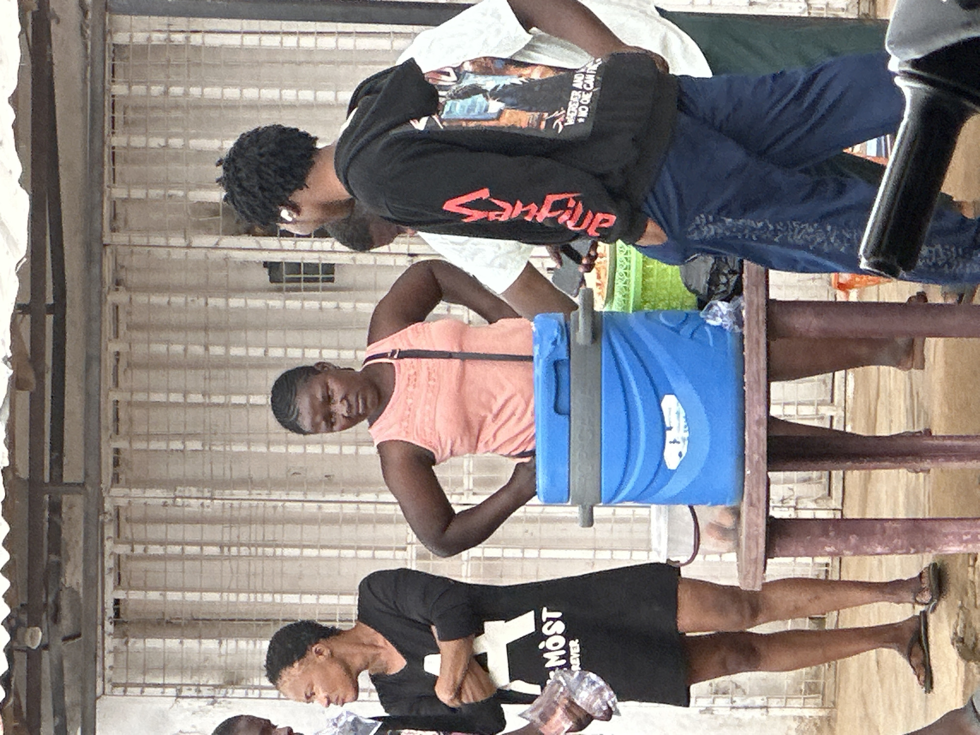 In Narayi, Nigeria, a group of people are gathered outdoors near a building with a corrugated metal roof and a white-paneled facade. In the foreground, a person wearing a black dress with a white "A MOST REVER" graphic on it stands with their arms crossed. To their right, a woman in a peach-colored tank top is holding her hands on her hips. Between them, a blue cooler sits on a round, weathered table. Behind these individuals, a young man in a black hooded sweatshirt with red lettering on the sleeve and a graphic on the back is visible, with his hand in his pocket. Another person in a white t-shirt and dark green garment stands behind him. The ground is a sandy or dusty earth, and what appears to be a motorcycle handlebar is in the extreme lower right corner of the frame.