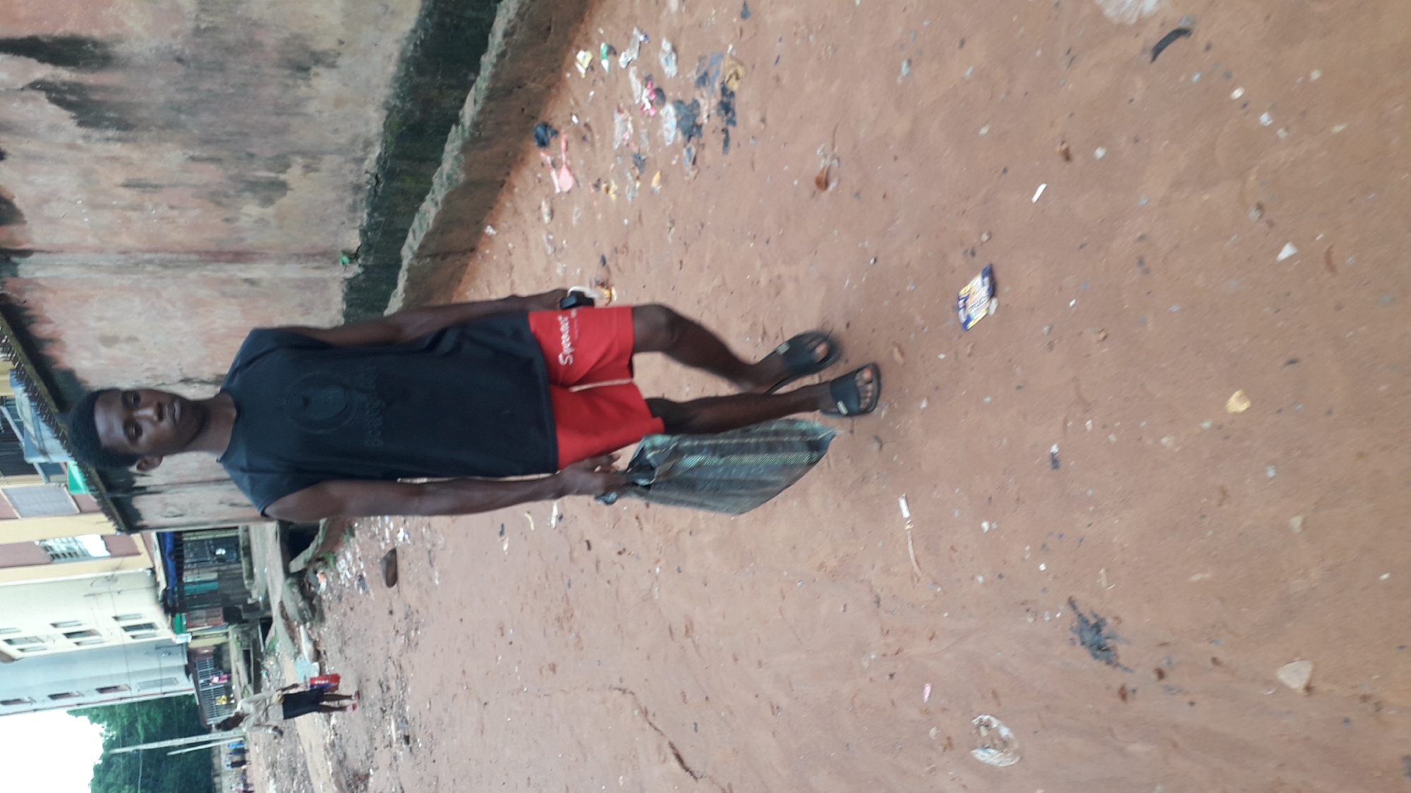 A young man stands on a dirt road in Onitsha, Nigeria. He wears a black t-shirt, red shorts with the word "Speas" printed on them, and black sandals. He holds a bag in his right hand. The road is littered with trash. In the background are buildings and other people. The scene appears to be an urban setting with signs of poverty and litter. A drainage channel is visible along the wall.