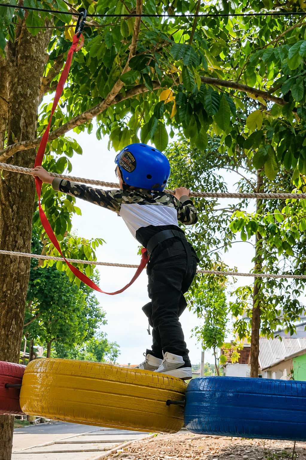 A child wearing a blue helmet and a camouflage jacket is navigating an obstacle course. The child is on a yellow tire suspended by ropes, with a red safety strap attached to their harness. The background shows lush green trees and a bright sky, suggesting an outdoor setting. The obstacle course appears to be set up in an area with some buildings visible in the distance.