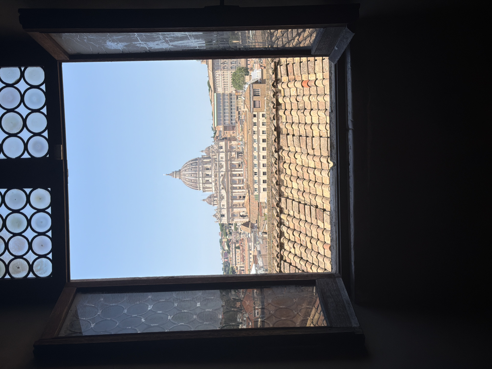 The image captures a view through an open window, looking out over a cityscape. The window frame is made of dark wood, with a patterned glass insert at the top featuring circular panes. The lower part of the window is open, revealing a terracotta-tiled rooftop in the foreground. Beyond the rooftop, a dense urban landscape unfolds, with numerous buildings of varying sizes and architectural styles. Dominating the background is a large, prominent building with a grand dome, characteristic of St. Peter's Basilica in Vatican City. The sky is clear and blue. The scene is illuminated by bright daylight, casting shadows within the room from which the photograph is taken.