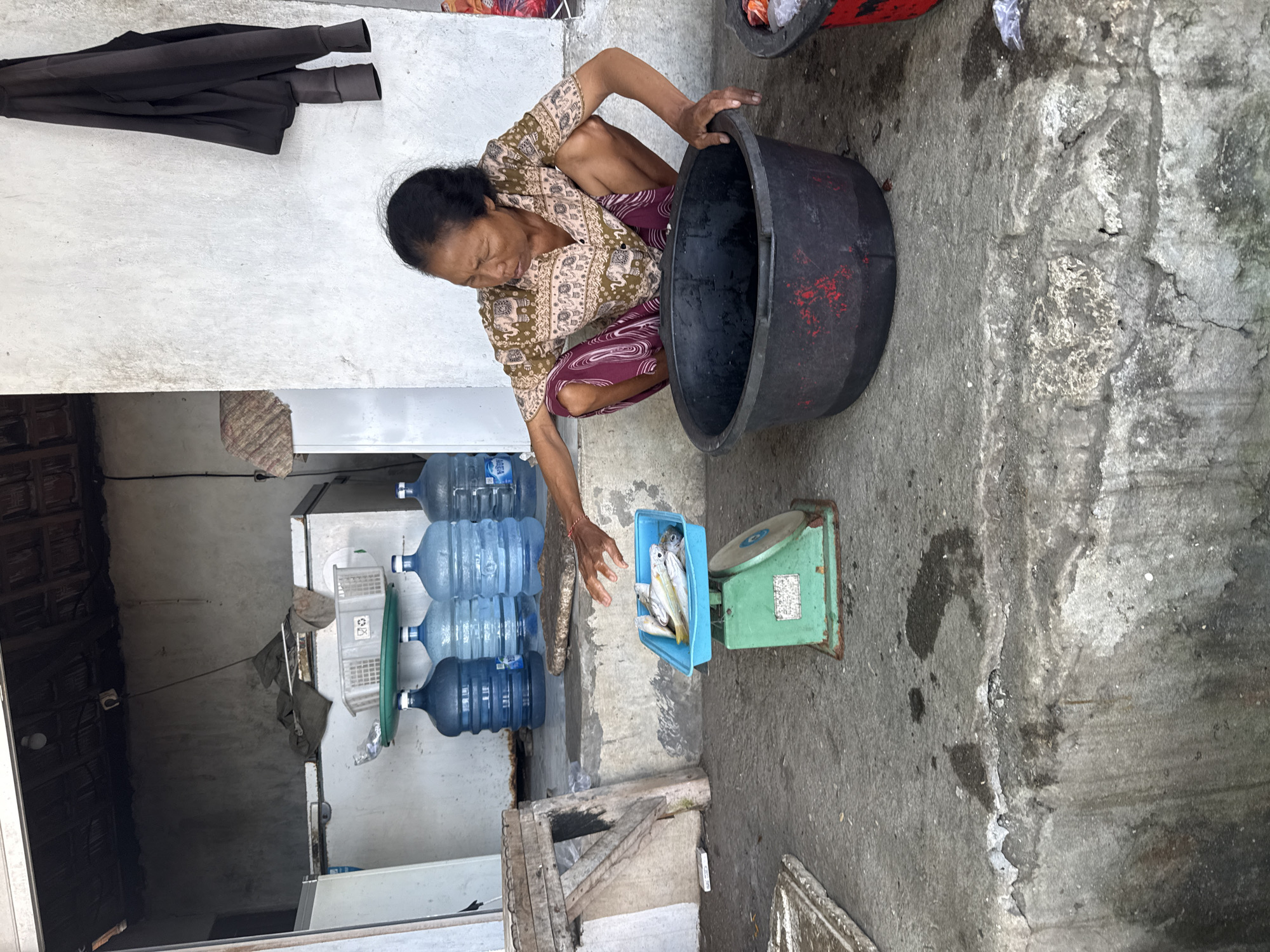 An Indonesian woman squats on the floor in Batununggal, Indonesia, holding a large, black, plastic tub over a green metal scale holding a blue plastic container filled with fish. Four large jugs of water sit against a wall in the background, beneath a concrete overhang. A black jacket hangs on the wall. The floor is concrete.