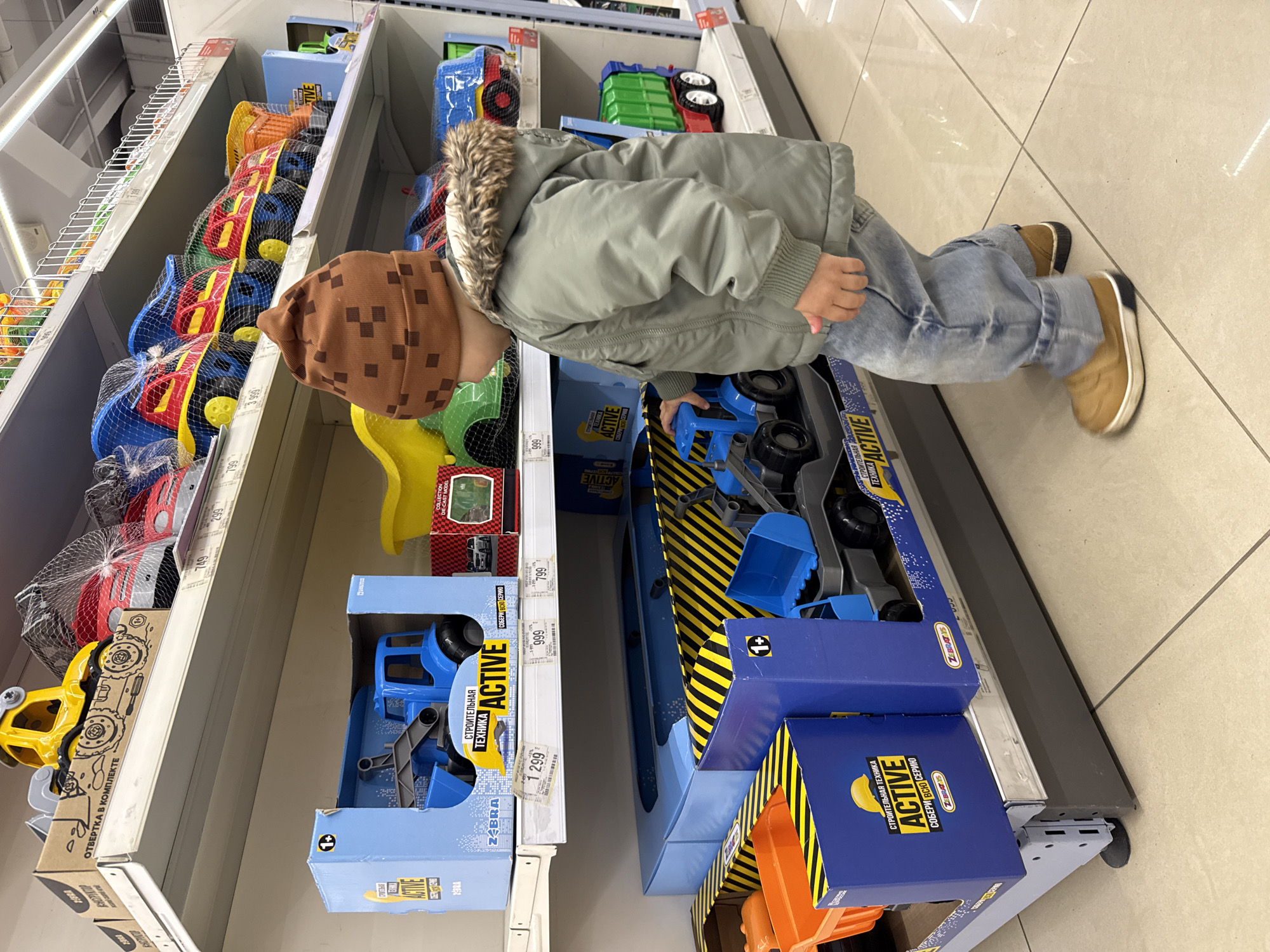 A young child wearing a brown checkered hat, a green jacket with a fur-lined hood, and blue jeans stands in front of a shelf in what appears to be a retail store, likely in the city of Korobovo, Russia, based on the presence of Russian language text on the product packaging. The child is leaning over and reaching into a box containing blue toy construction vehicles. The shelf is stocked with various other toys, including plastic vehicles in nets and boxed sets with the words "СТРОИТЕЛЬНАЯ ТЕХНИКА ACTIVE" (Construction Equipment Active) prominently displayed in Russian. Price tags are visible on the shelf edges, indicating the cost of the items. The floor is light-colored tile. The overall scene suggests a consumer environment, specifically the toy section of a store.