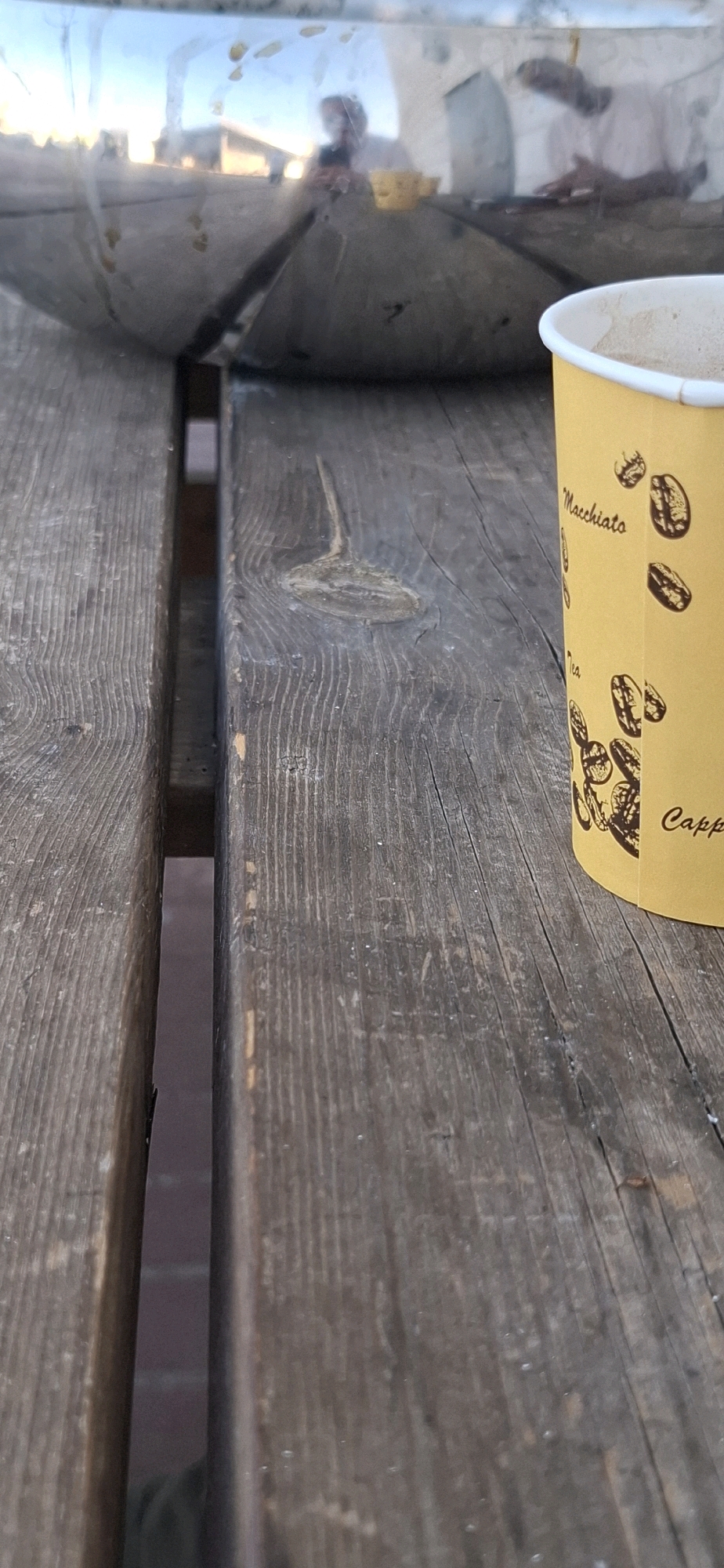 A partially filled yellow coffee cup sits on a weathered wooden picnic table. The cup has brown coffee bean illustrations and text that reads "Macchiato" and "Capp". In the background, a reflective surface, possibly a metal bowl, shows a distorted reflection of a person holding a phone and another person with their hands on a table. The environment suggests an outdoor seating area. The image was taken in Kiryat Gat, Israel.
