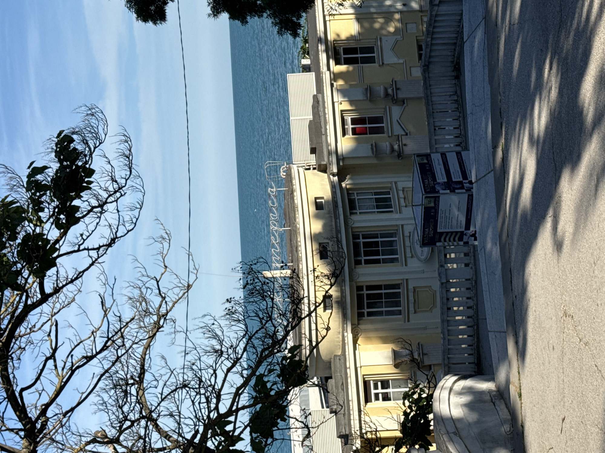 This image depicts a street scene with a building and a body of water. The perspective is from a low angle, looking upwards, and the image is rotated 90 degrees counter-clockwise, making the scene appear sideways.
In the foreground, bare tree branches dominate the left side of the frame, silhouetted against a bright blue sky with wispy white clouds. The sky suggests it is daytime, likely morning or afternoon given the clarity and brightness.
Behind the trees, a multi-story building with a light yellow facade is visible. It has numerous windows, some with white frames and others with red curtains. The building has decorative architectural elements, including ornate carvings around some windows and decorative urns on columns. A white railing runs along a balcony or walkway in front of the building.
To the left of the building, a vast expanse of blue water, possibly the sea or a large lake, stretches to the horizon under the blue sky.
A grey paved walkway or sidewalk is present in front of the building, casting dappled shadows from unseen trees. On the sidewalk, there are several upright signs with text. One sign clearly shows the words "VERONA SUMMER" and another has the number "100". The text suggests a summer event or festival in Verona.
The overall impression is a calm, possibly vacation-like setting, with the juxtaposition of urban architecture, natural elements like trees and water, and promotional signage. There are no visible people in the frame. The lighting suggests a sunny day.