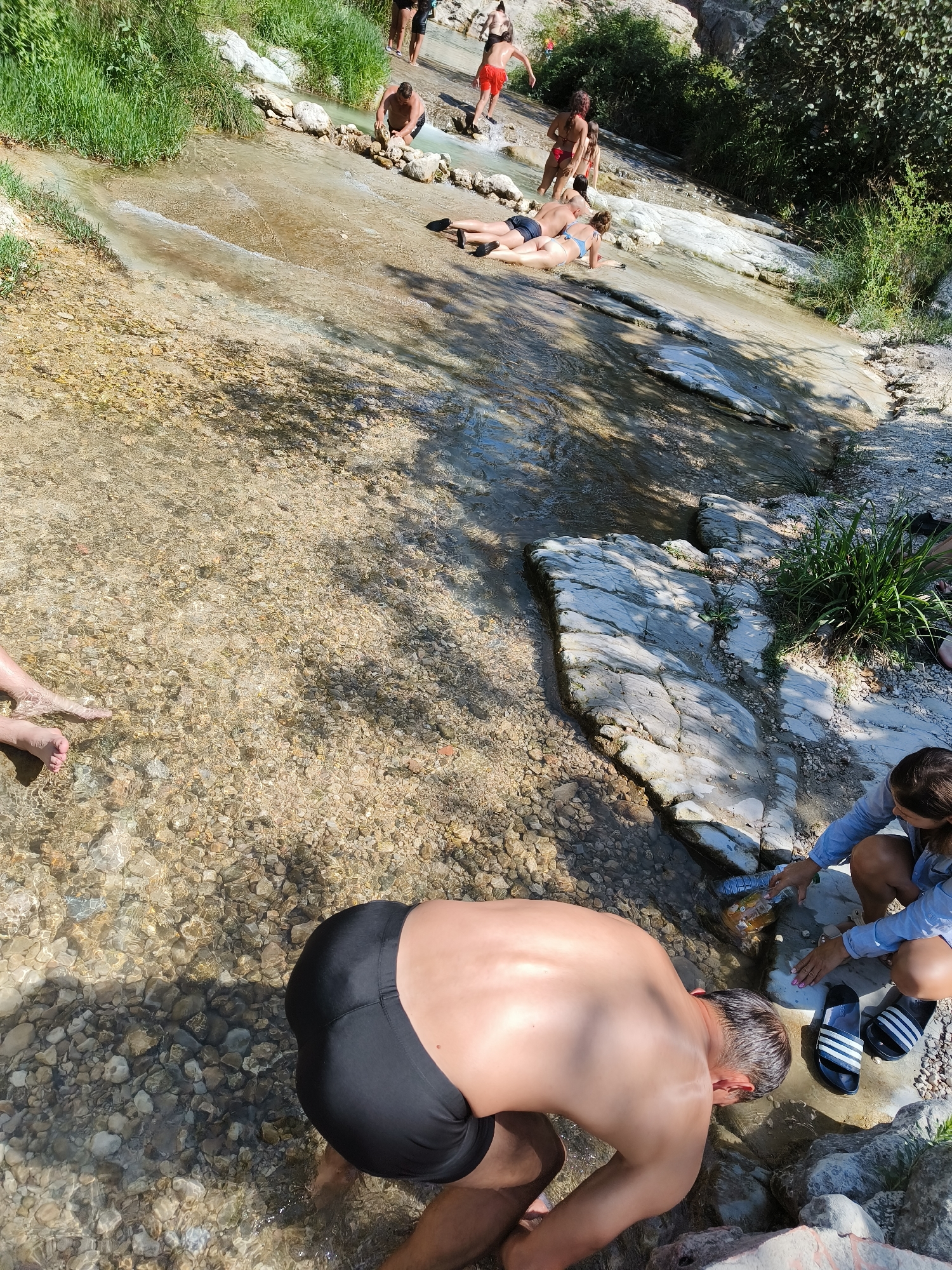 This image depicts a group of people bathing in a shallow, rocky stream with a gentle current. The stream appears to be a natural watercourse flowing over light-colored, smooth stones and gravel. Lush green vegetation lines the banks of the stream, and some of the rocks are covered in moss. In the background, there are indications of a natural, possibly mountainous, landscape.
In the foreground, a man wearing black swim trunks is bending over, apparently washing his hands or face in the water. To his right, a woman in a blue shirt and dark shorts is also interacting with the water, holding a plastic bottle. Next to her are a pair of black and white striped slides. Further upstream, several other individuals are visible, some standing in the water and others lounging on the rocks. One person is wearing red swim trunks, another is in a bikini, and a couple is lying down on the rocks near the water's edge. The overall impression is of a relaxed, outdoor recreational activity in a natural setting.
The location is identified as the Pou Clar natural pools in Ontinyent, Spain. These pools are known for their clear water, sculpted rock formations, and surrounding natural beauty, making them a popular spot for swimming and picnicking during warmer months.