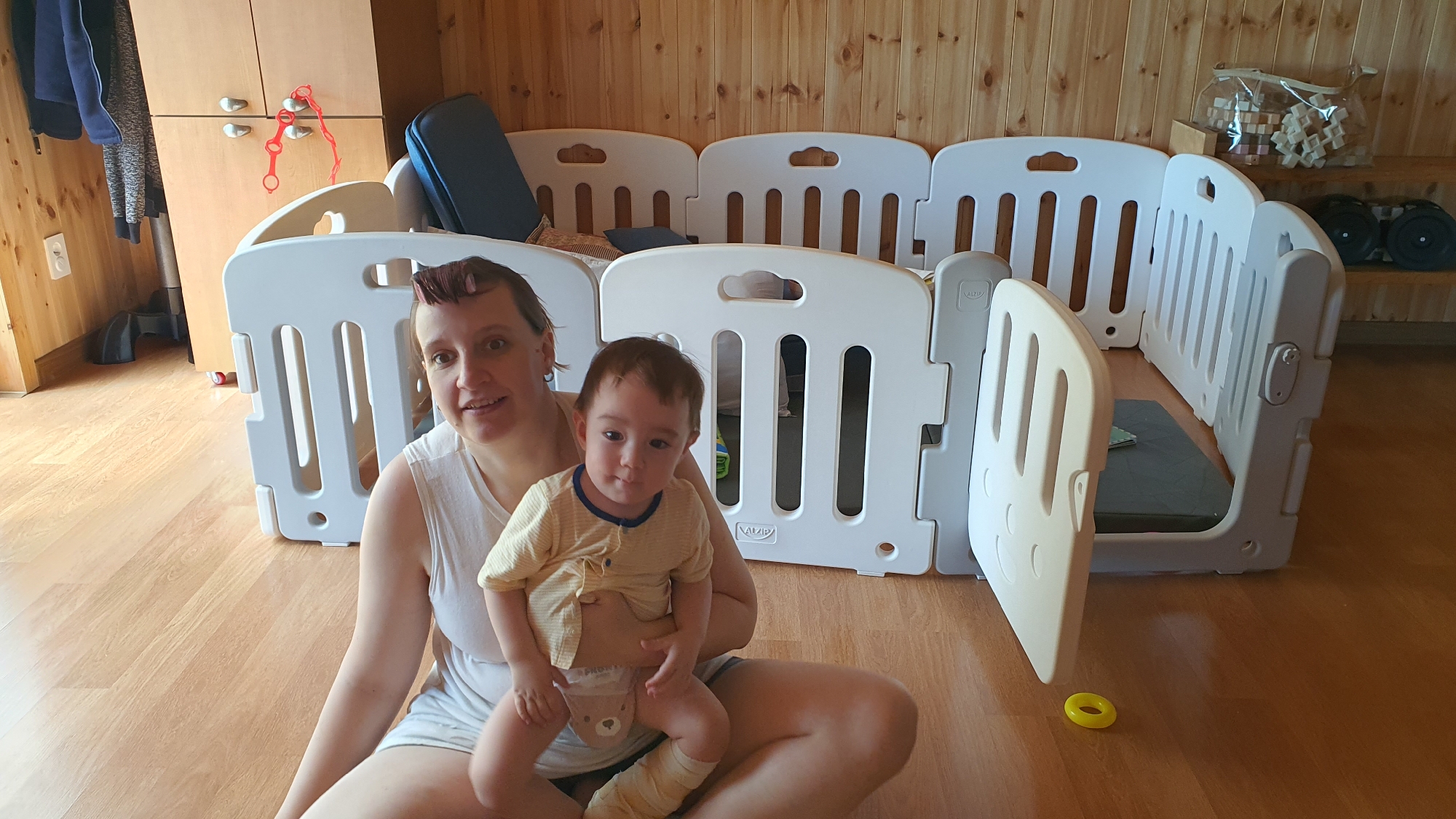 A woman sits on a wooden floor holding a baby in front of a white plastic playpen. The playpen encloses a gray mat. A yellow ring toy lies on the floor. A wooden cabinet is visible in the background, along with wood-paneled walls and a shelf with toys and weights. The location appears to be inside a home.