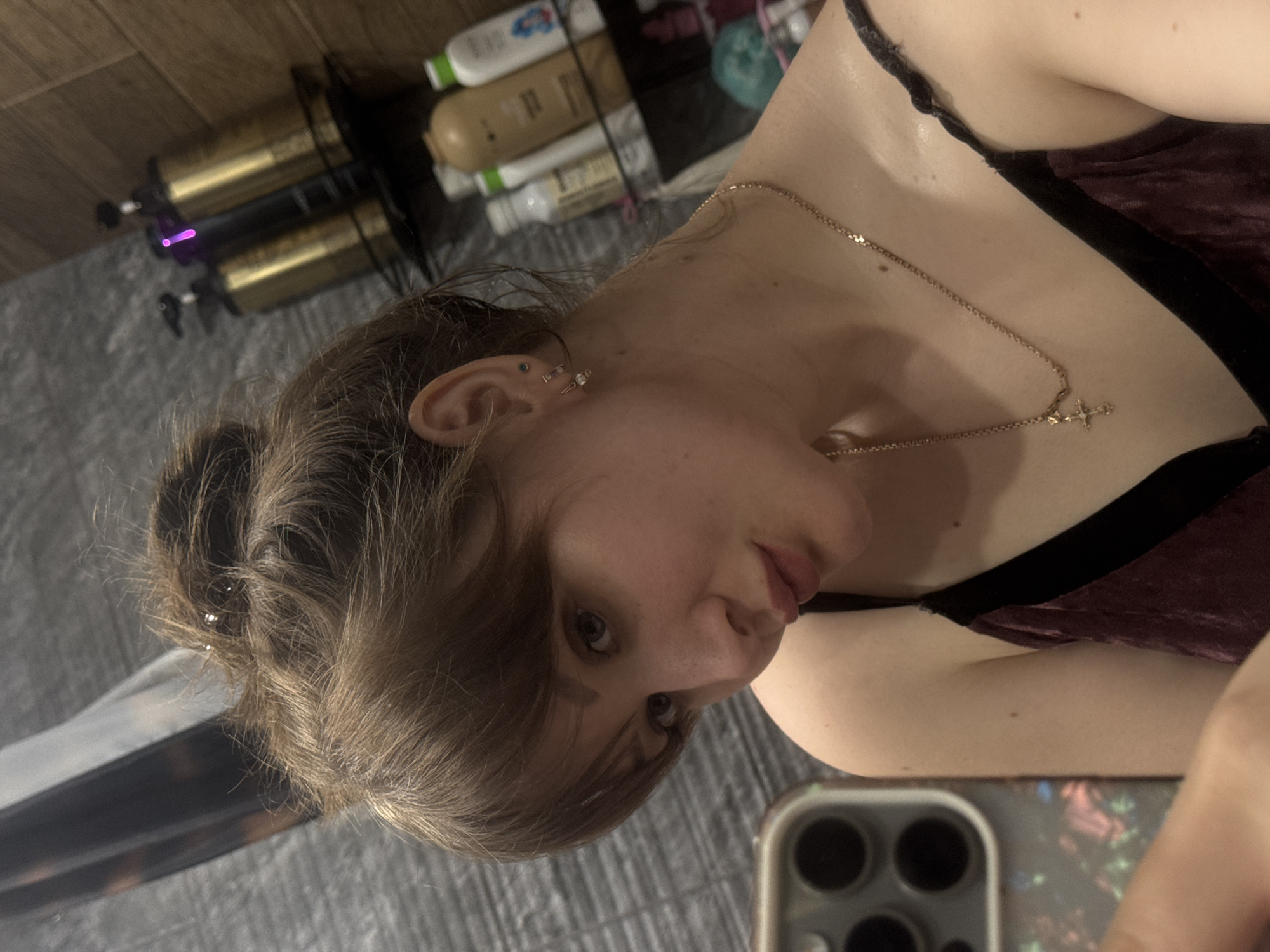 A person with fair skin and light brown hair is posing for a selfie. The hair is styled in a loose bun with some strands framing the face. The person is wearing a dark-colored velvet top with black straps and a delicate gold necklace with a small cross pendant. The background includes shelves with various bottles and containers, and a section of a gray bedspread or fabric. The phone being used to take the picture is visible in the lower right corner. The location is not discernible as being in Chaikovsky, Russia, based on the visual information provided.
