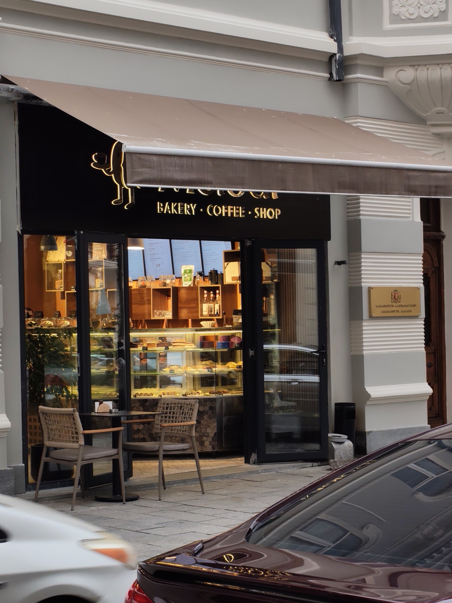 The image shows the exterior of a bakery and coffee shop in Batumi, Georgia. The shop has a large glass storefront displaying various pastries and baked goods. Two chairs and a small table are set up outside the shop on the sidewalk. The shop's signage, illuminated in yellow neon, reads "BAKERY COFFEE SHOP." Above the entrance, an awning provides shade. A plaque with Georgian script and a coat of arms is affixed to the building's facade to the right of the shop entrance. Two cars are partially visible in the foreground, one white and one dark red, suggesting the shop is located on a street. The architecture of the building is classic, with decorative moldings. The overall scene depicts a typical urban street view in Batumi.