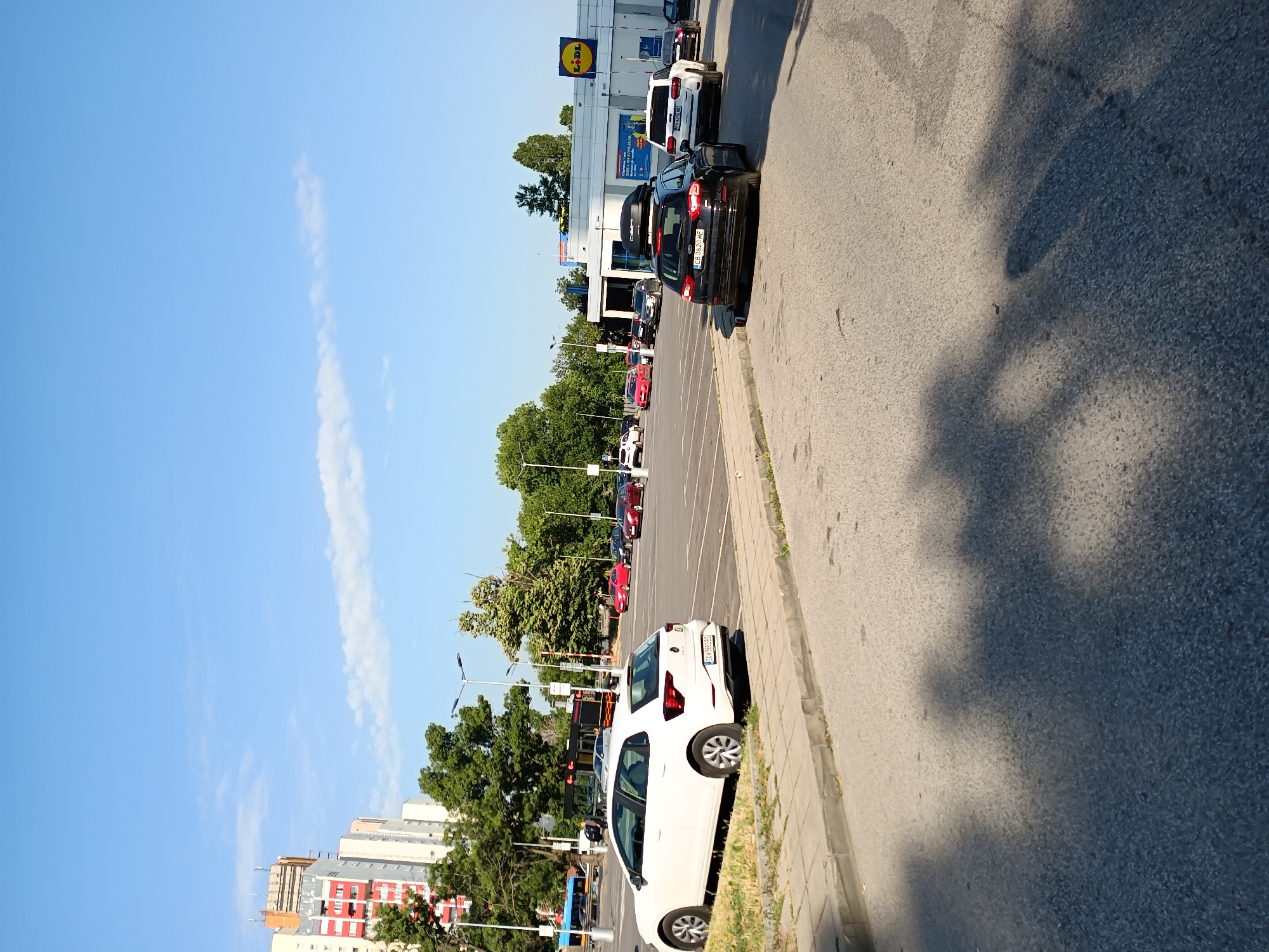 The image depicts a parking lot in front of a Lidl supermarket in Sofia, Bulgaria. Several cars are parked in the lot, including a white Volkswagen Polo in the foreground. In the background, there are tall apartment buildings and trees. The sky is blue with a few scattered clouds. The scene appears to be a typical day in a commercial area of the city.