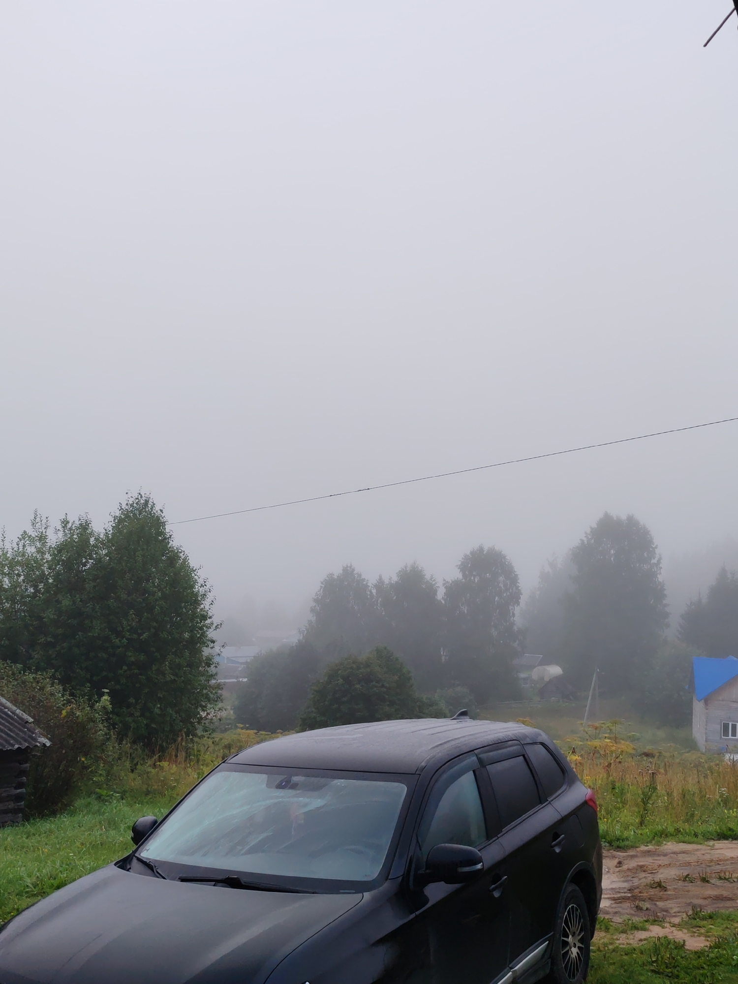 The image depicts a rural scene in the city of Chukhlem, Russia, on a foggy day. In the foreground, a black SUV is parked on a dirt and grass track. The vehicle's windshield is partially obscured by condensation or rain. Behind the SUV, a landscape unfolds with lush green trees and vegetation, though visibility is reduced by a thick fog that obscures the distant background. Several rural structures, including a small wooden building with a corrugated roof to the left and a larger house with a blue roof to the right, are visible amidst the trees and fog. A thin power line stretches horizontally across the upper middle section of the frame. The overall atmosphere is one of quiet, overcast weather in a remote, possibly village setting.