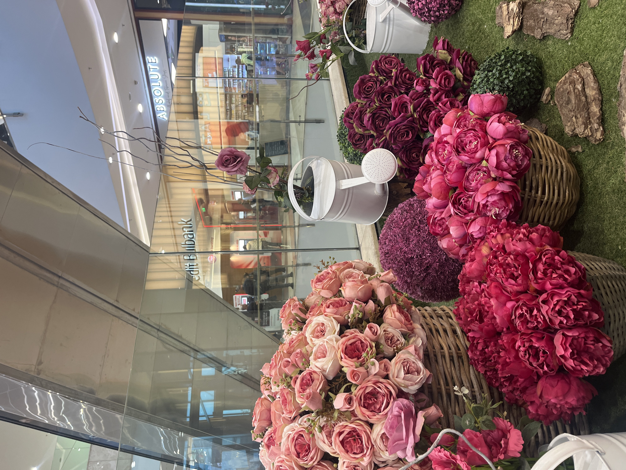 The image shows a floral display in a shopping mall setting. In the foreground, there are baskets filled with various types of flowers, including pink roses and dark red peonies. Two white watering cans are integrated into the display. The background features the glass storefronts of shops. Signs for "Absolute" and "Credit Bulbank" are visible. The location is likely in Sofia, Bulgaria, due to the presence of Credit Bulbank, a Bulgarian bank.