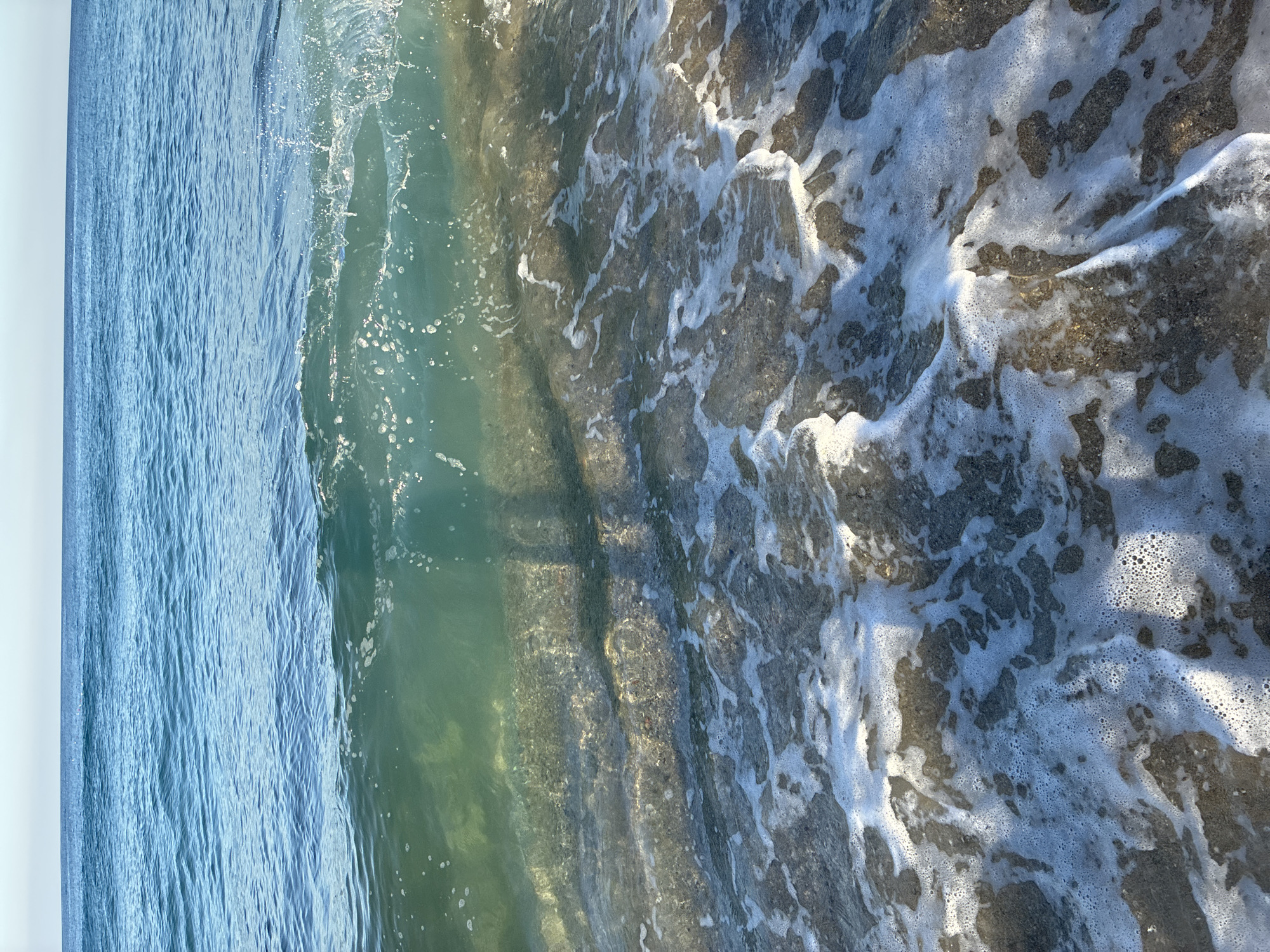 The image depicts a close-up view of a wave breaking on a sandy beach in Alanya, Turkey. The water is clear and turquoise, with sunlight illuminating the sandy bottom beneath the surface. Foam from the breaking wave is spread across the sand, creating a frothy texture. In the distance, the calm blue sea stretches towards the horizon under a clear sky. A faint shadow of a person is visible within the wave, suggesting the presence of a photographer or observer. The scene captures a typical moment of a sunny day at the beach.